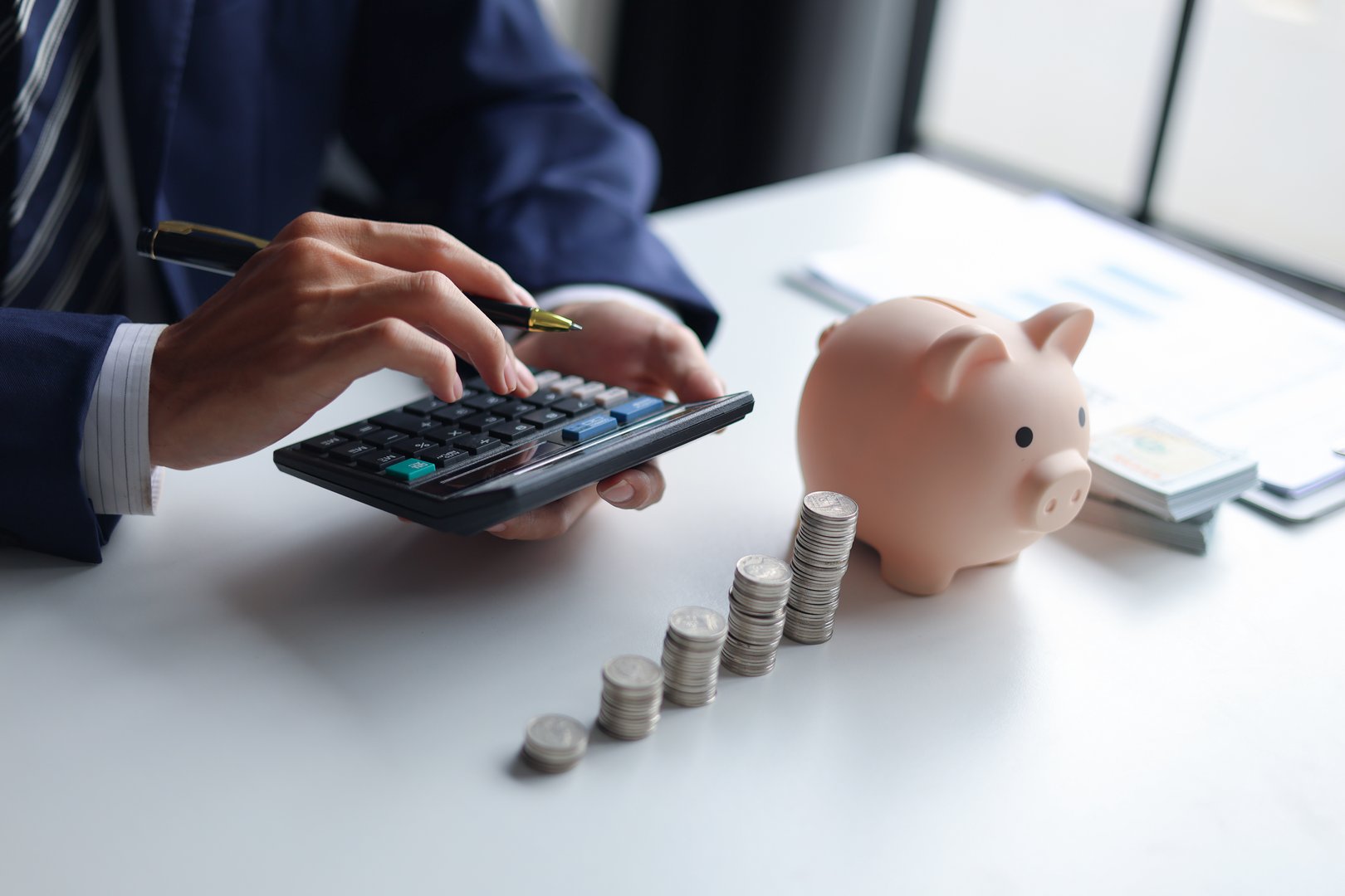 A person using a calculator with a piggy bank, stacks of coins, and US dollar bills on a table. Concept of saving money and financial planning.