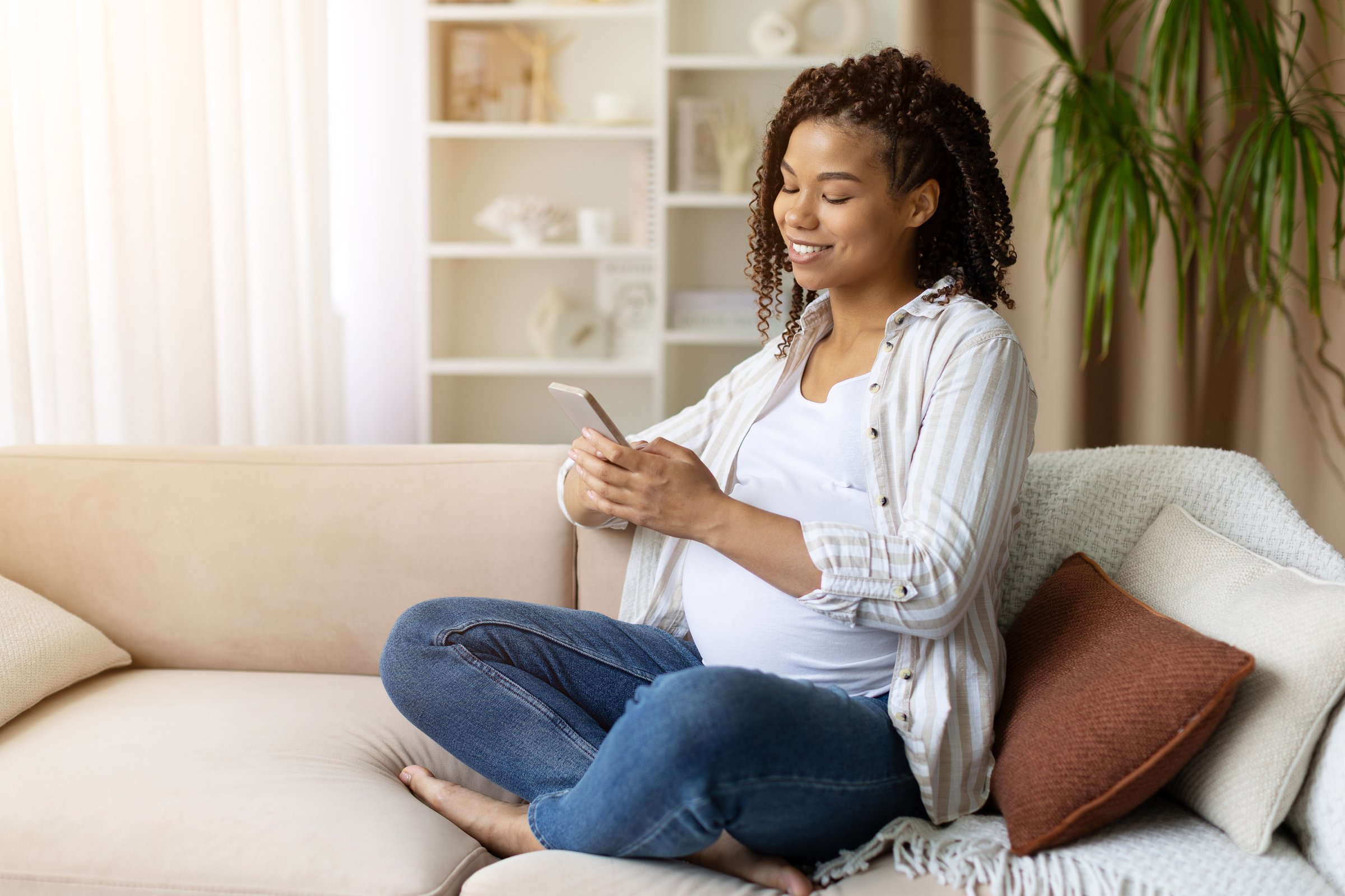 Smiling young black pregnant woman sits cross legged on comfy sofa using smartphone at home. Casual jeans and shirt, relaxed mood. Concept of prenatal care, communication, lifestyle balance