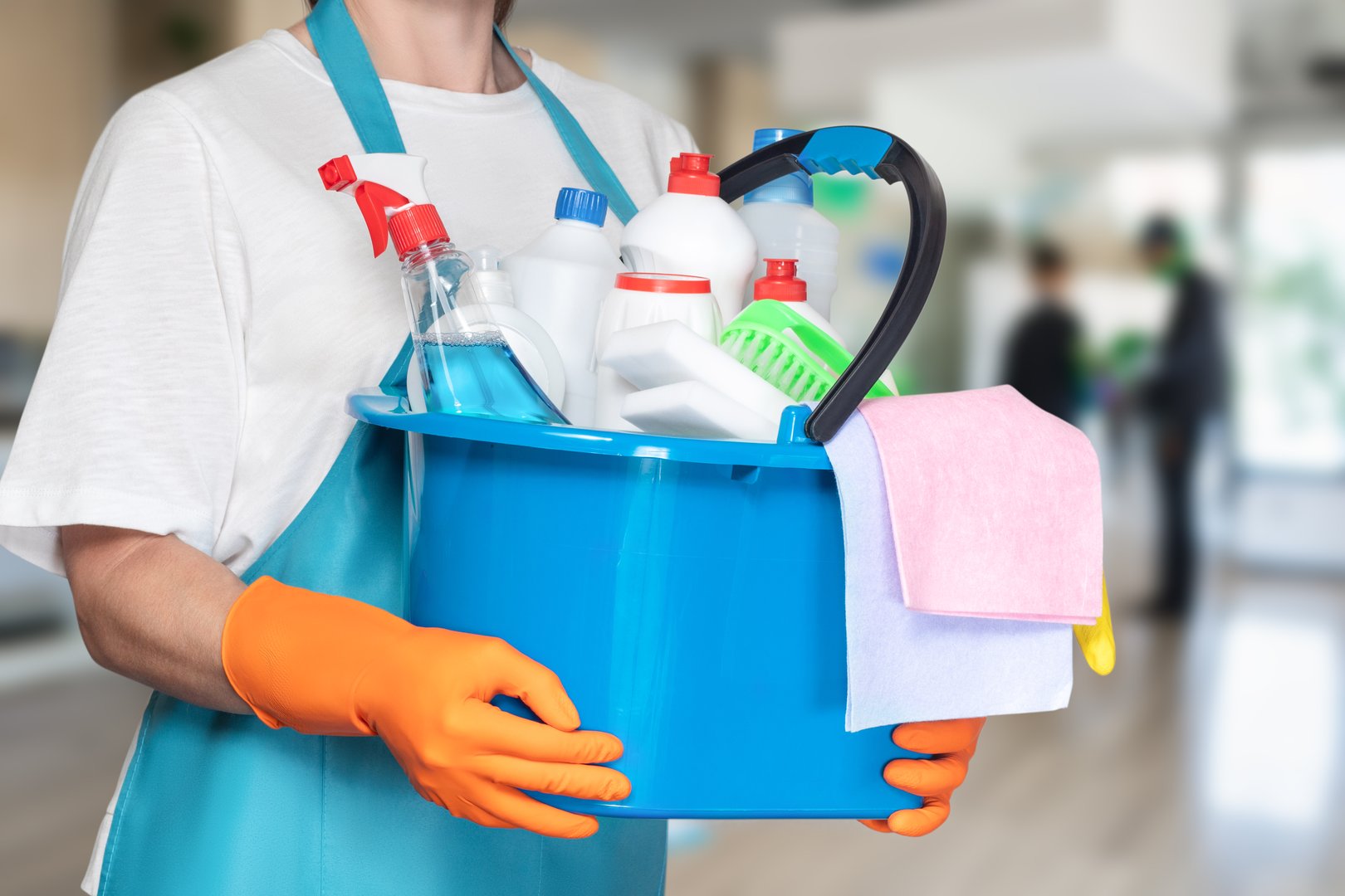A person wearing gloves holds a bright blue bucket filled with various cleaning supplies. The setting appears to be an indoor area, ready for a thorough cleaning session.