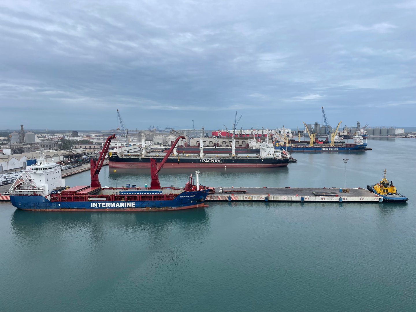 Cargo ships and cranes in a busy port under an overcast sky.