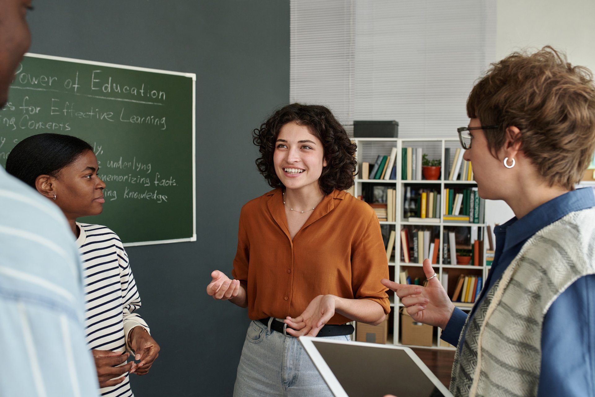 Diverse group of adults engaged in discussing educational concepts in casual office setting, with chalkboard in background showing informational notes