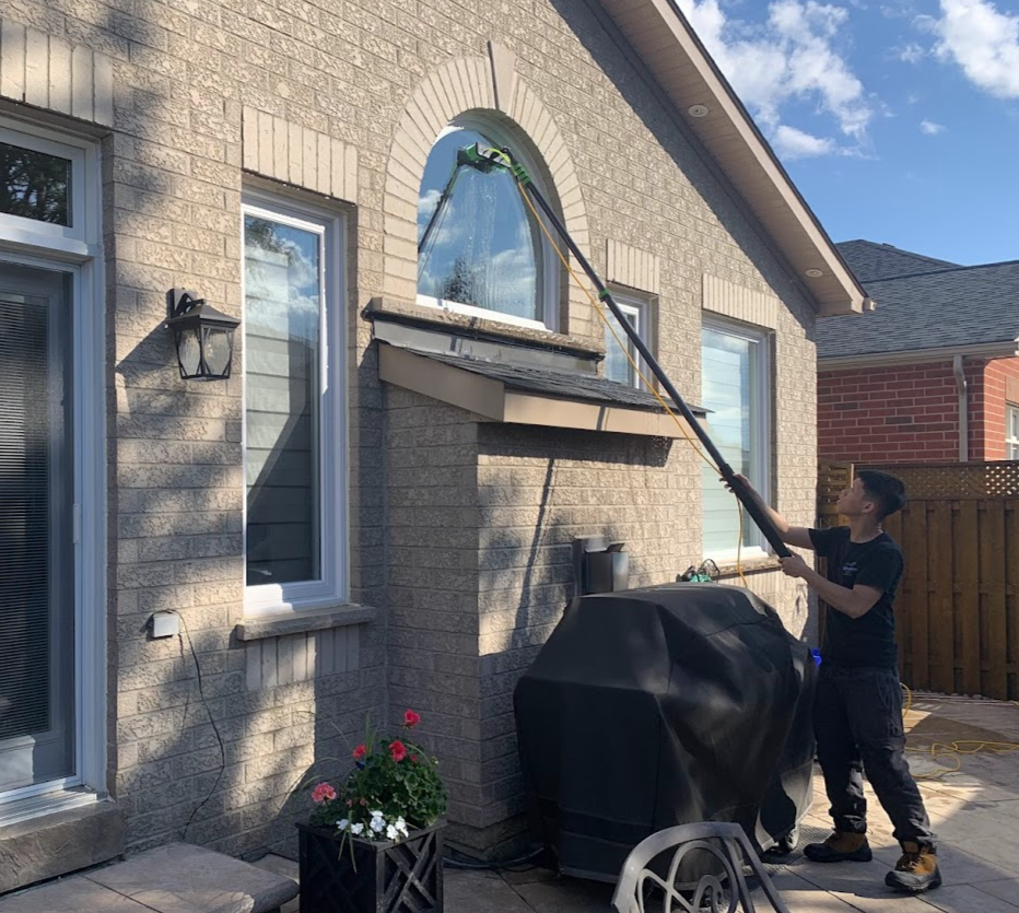 Person cleaning an arched window on a brick house using an extendable pole beside a patio with potted flowers.