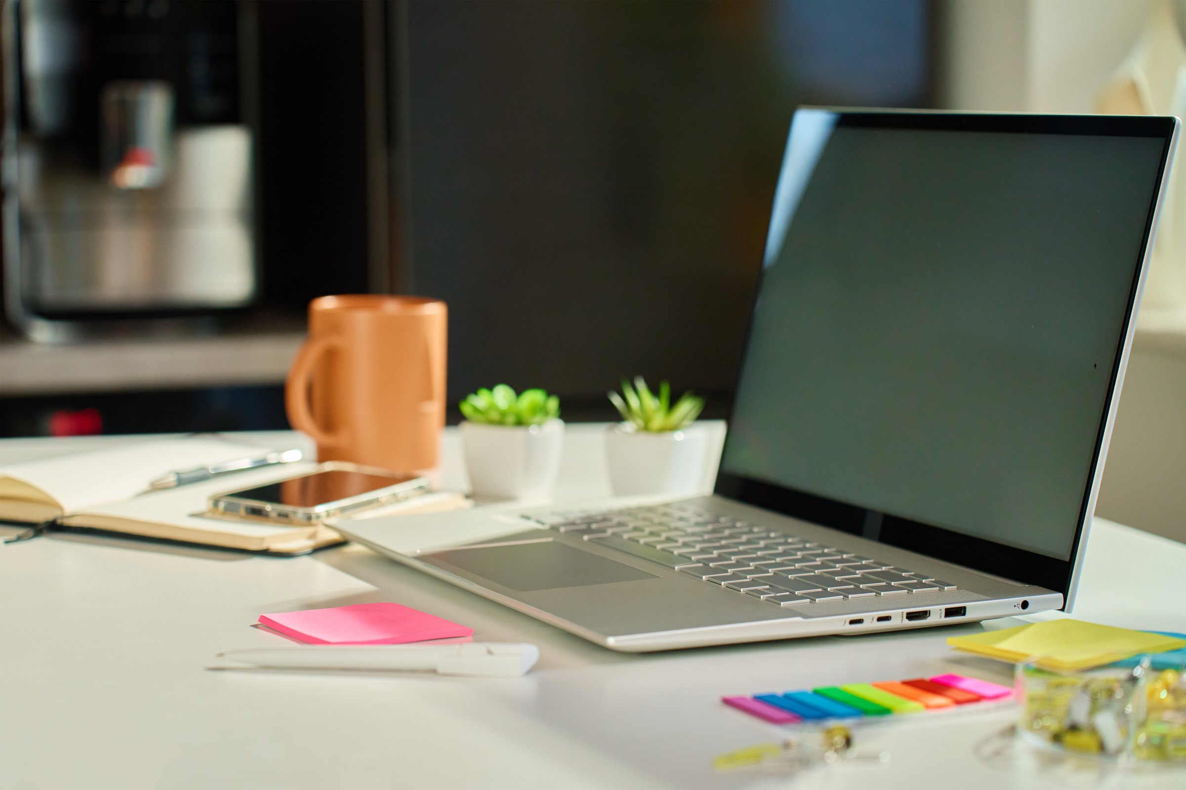 Minimalist home office with laptop, small potted plants and coffee mug on white desk with kitchen interior in background. Workspace for distant work from home