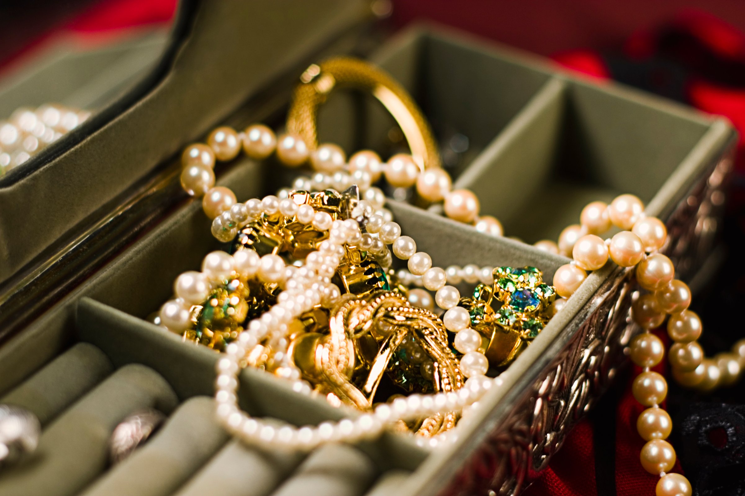 An open jewelry box with pearl necklaces, gold bracelets, and gemstone earrings inside, set against a dark background.