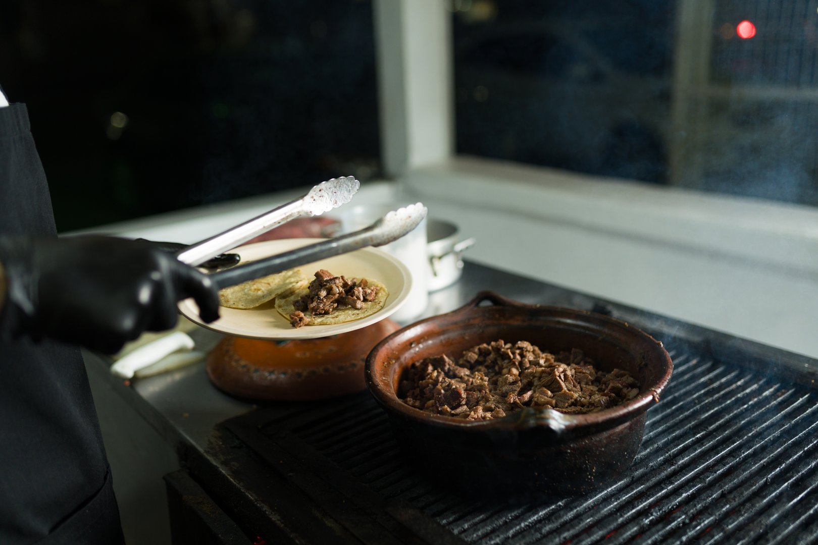 Chef using tongs to serve meat on tortillas, cooking tacos in a traditional mexican taqueria