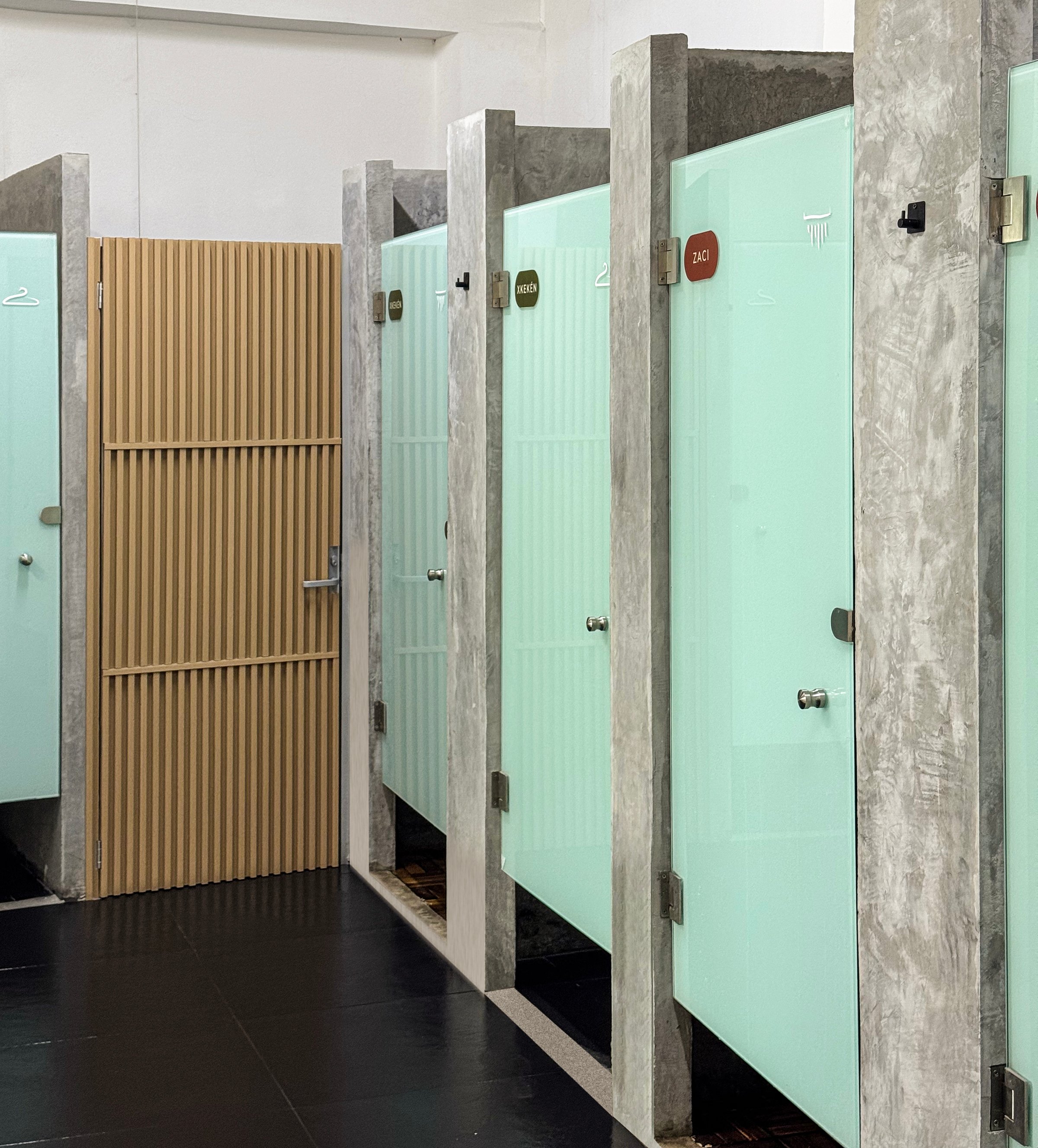 A row of modern bathroom stalls with frosted glass doors and concrete dividers, next to a ribbed wooden door.