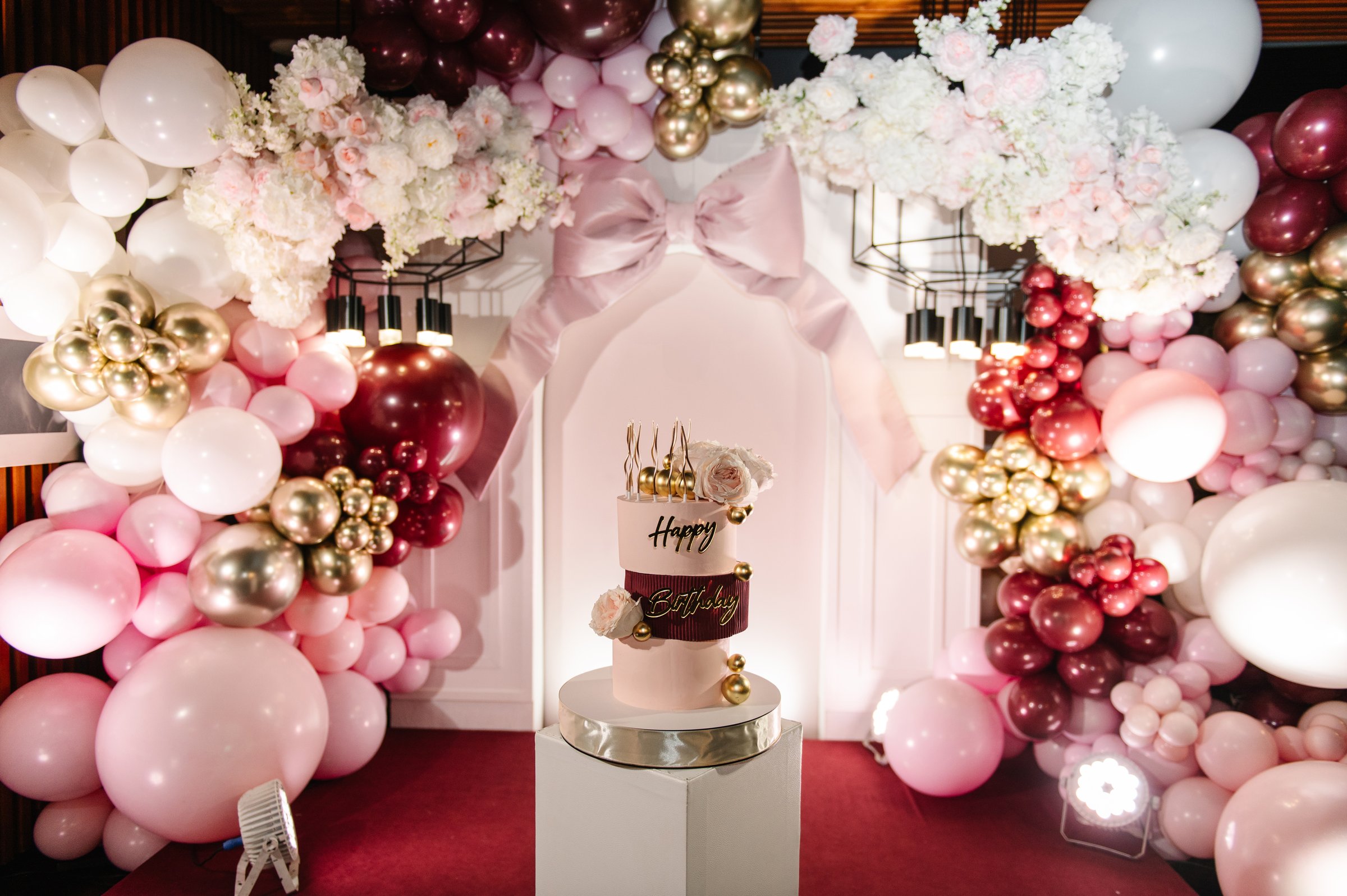 Trendy burgundy Cake with candles on background balloon and big bow on arch. Celebration concept. Birthday party decorations. Three-tiered cake with pink roses, Happy Birthday text topper, gold decor.