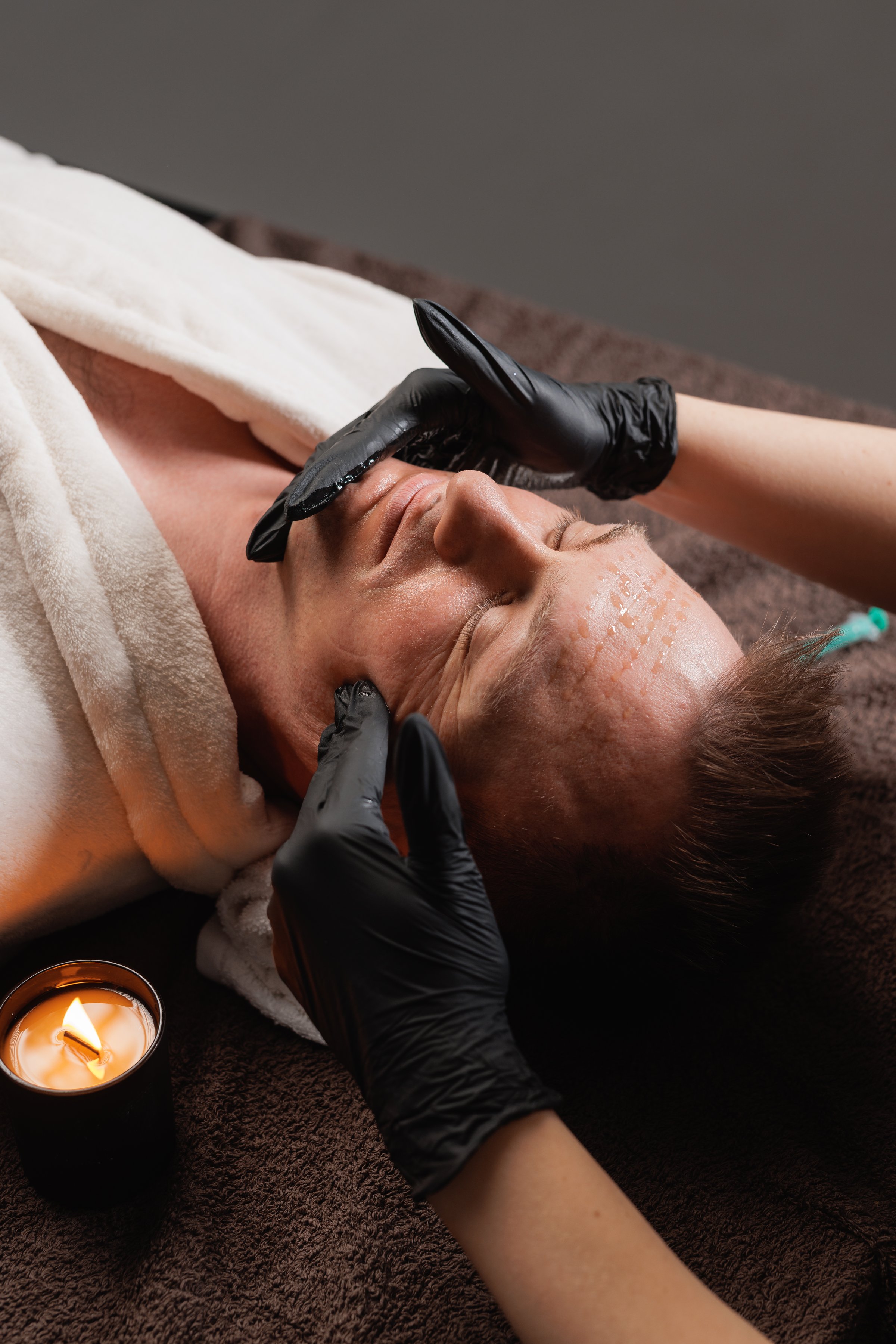 Adult man in bathrobe receiving relaxing facial massage after mesotherapy in spa clinic, concept of male antiaging skincare and recovery.
