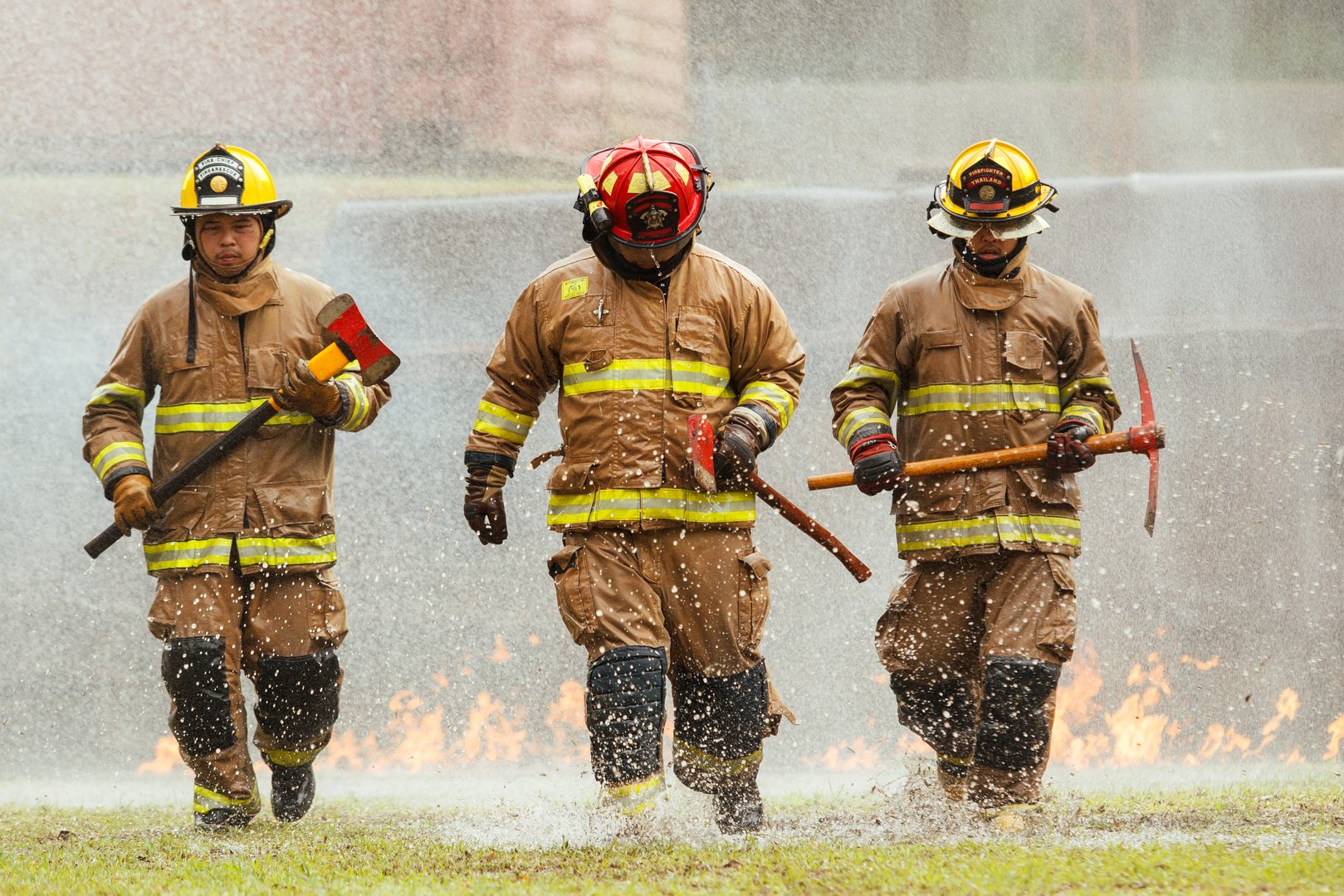 Team of firefighters in full protective gear carrying axes and tools while walking through water and flames during emergency training. Concept of teamwork, rescue operation, safety, and disaster response.