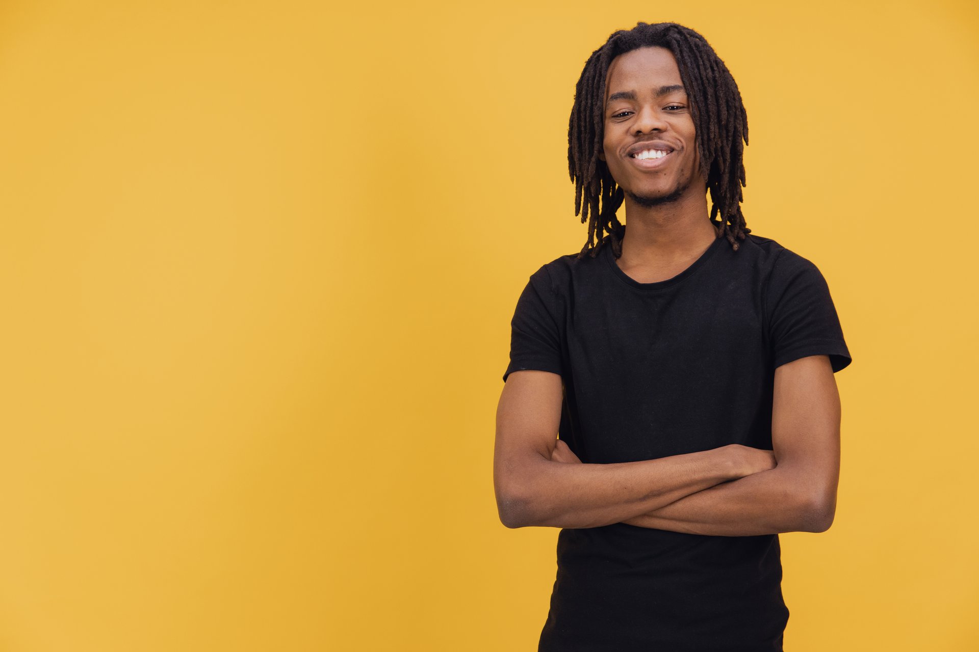 Portrait of a cheerful young African American man with dreadlocks smiling with crossed arms on a vibrant yellow background, exuding confidence and positivity