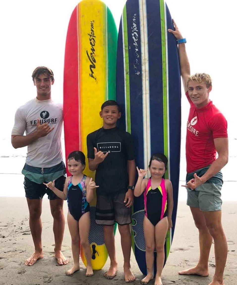 Three surfers with colorful boards and two children pose on a beach, all making a shaka hand sign.