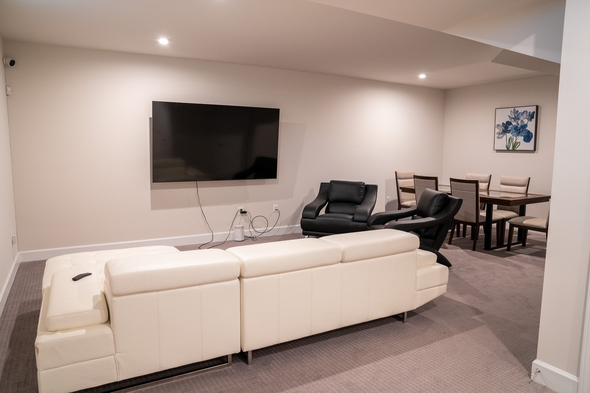 Modern living room with white sofa, wall-mounted TV, chairs, and a dining table. Neutral tones and minimal decor.