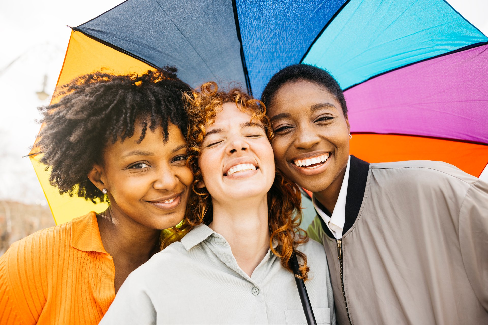 Three cheerful multi-ethnic young women friends are posing under a colorful rainbow umbrella