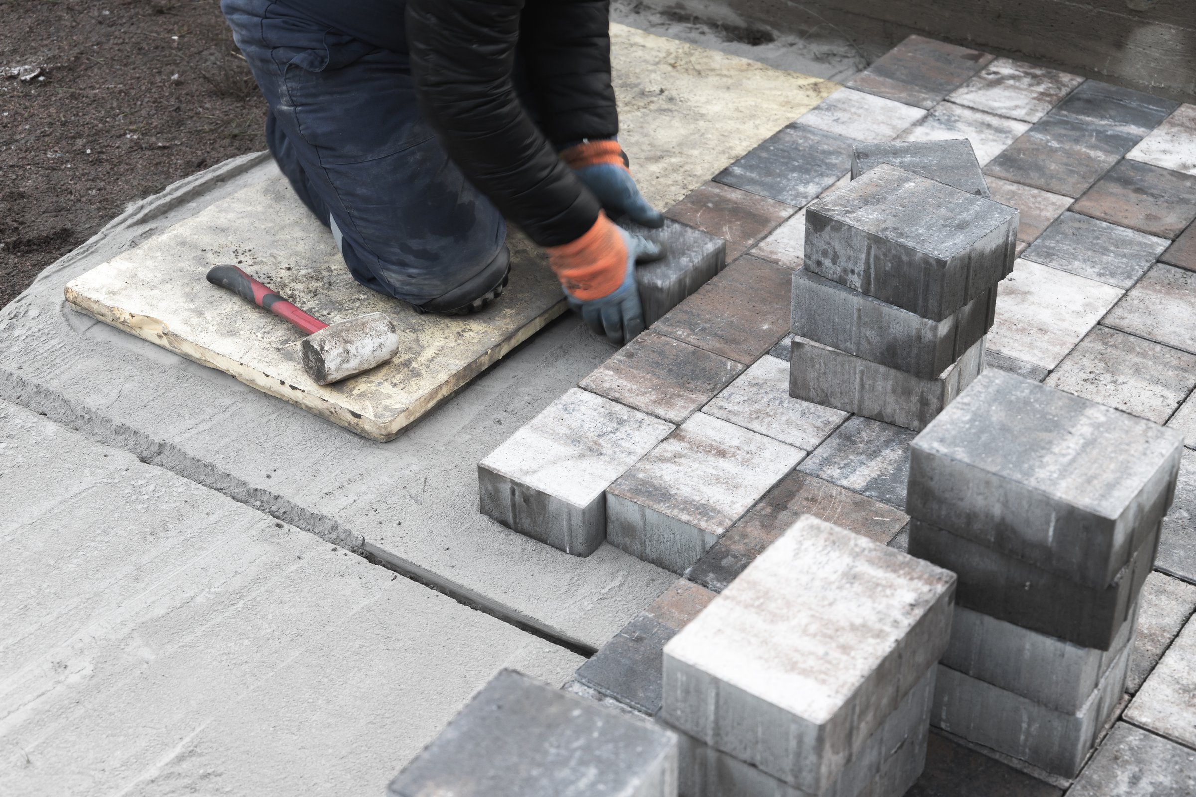 A skilled laborer carefully laying concrete bricks to create a paved surface outdoors, ensuring precision and durability in construction. Nearby, tools are arranged for use
