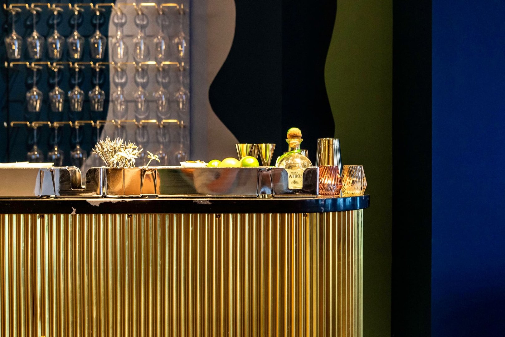 Luxurious bar setup with gold trim, glasses on a rack, limes, a bottle of liquor, and cocktail shakers.