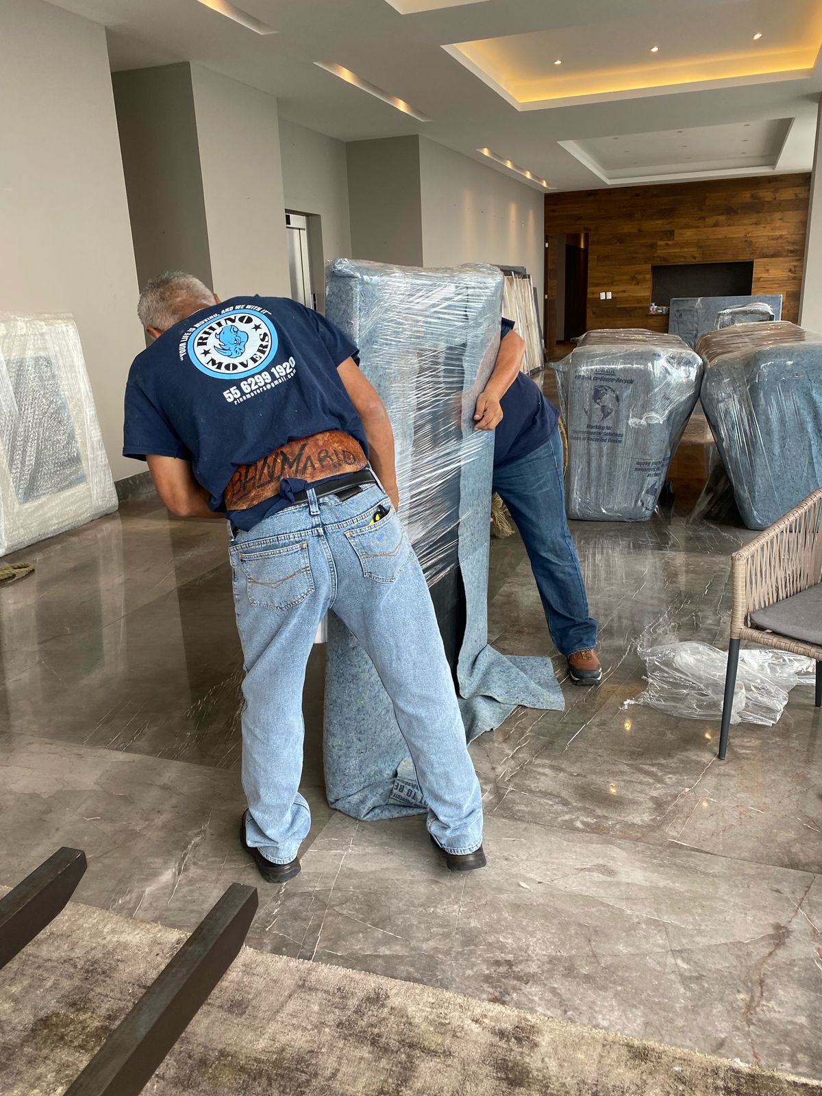 Two men unpacking furniture wrapped in plastic in a spacious room with a wooden accent wall and modern decor.