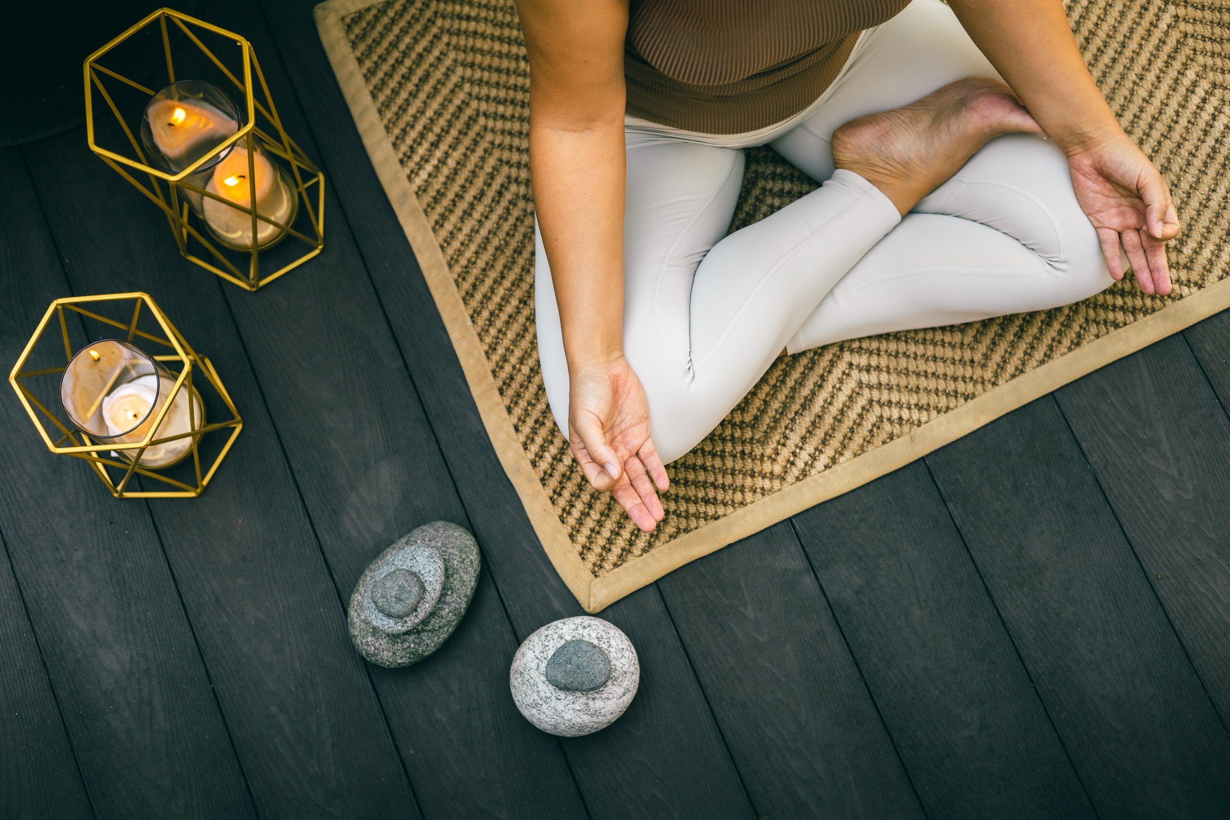 Close-up of a person sitting in lotus pose on a woven mat with hands resting on knees, surrounded by lit candles and stacked zen stones on a dark wooden floor in calm ambiance