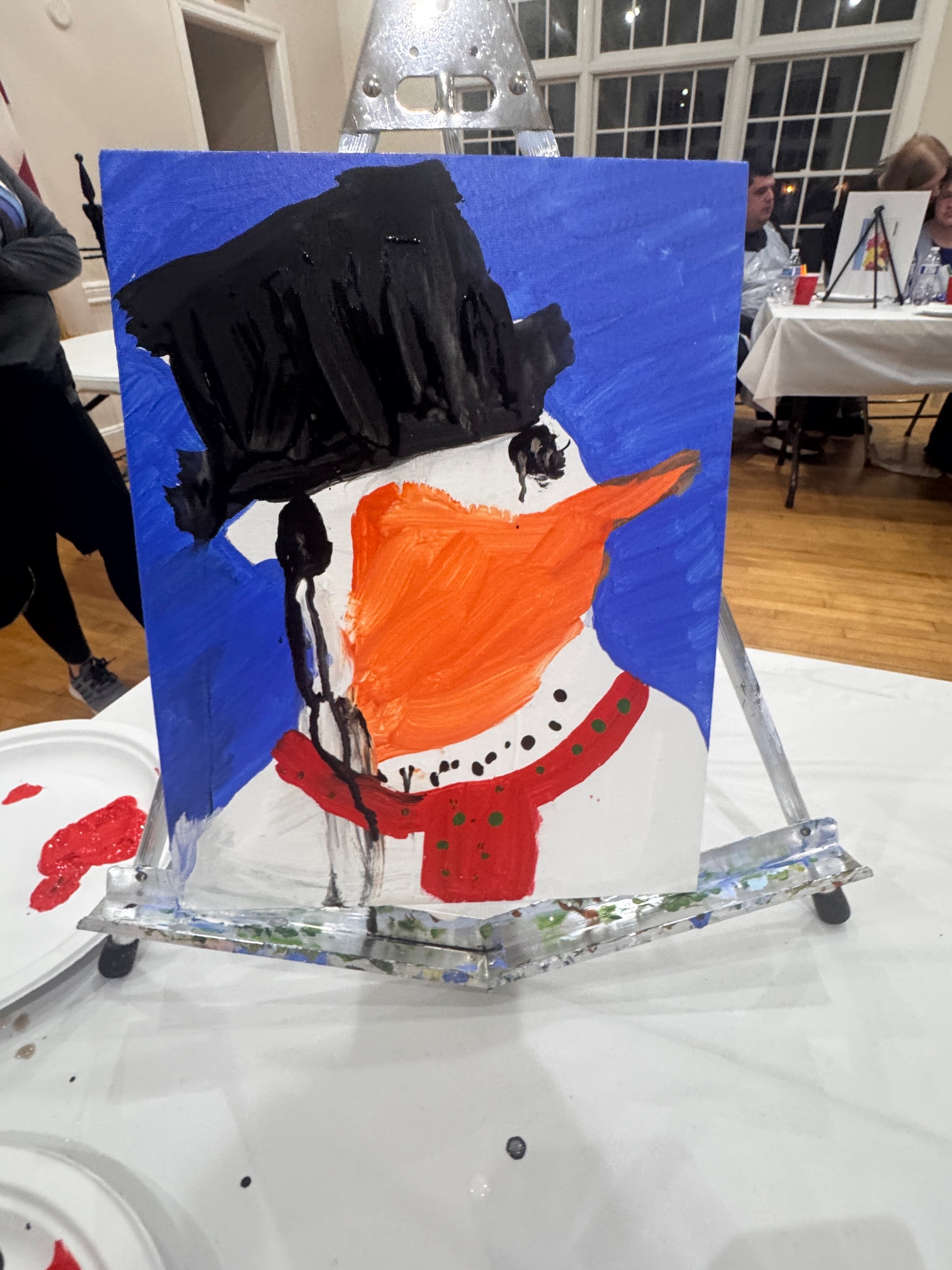 Painting of a snowman with a black hat, orange carrot nose, and red scarf on an easel in an art class setting.