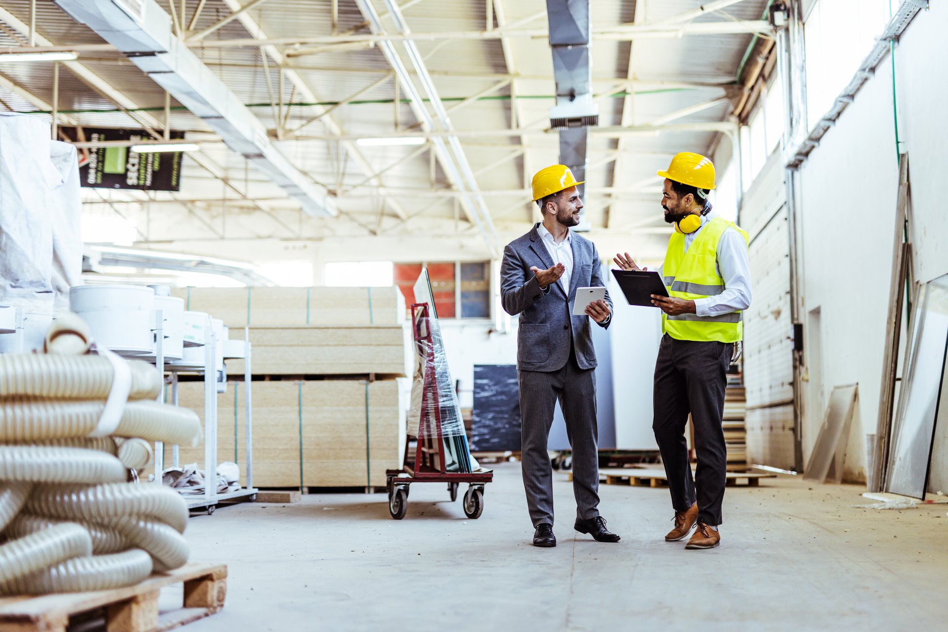 Industrial engineers discussing project details inside a large manufacturing facility, holding digital tools and equipment. Both wear protective gear and focus on teamwork and productivity in a professional environment.