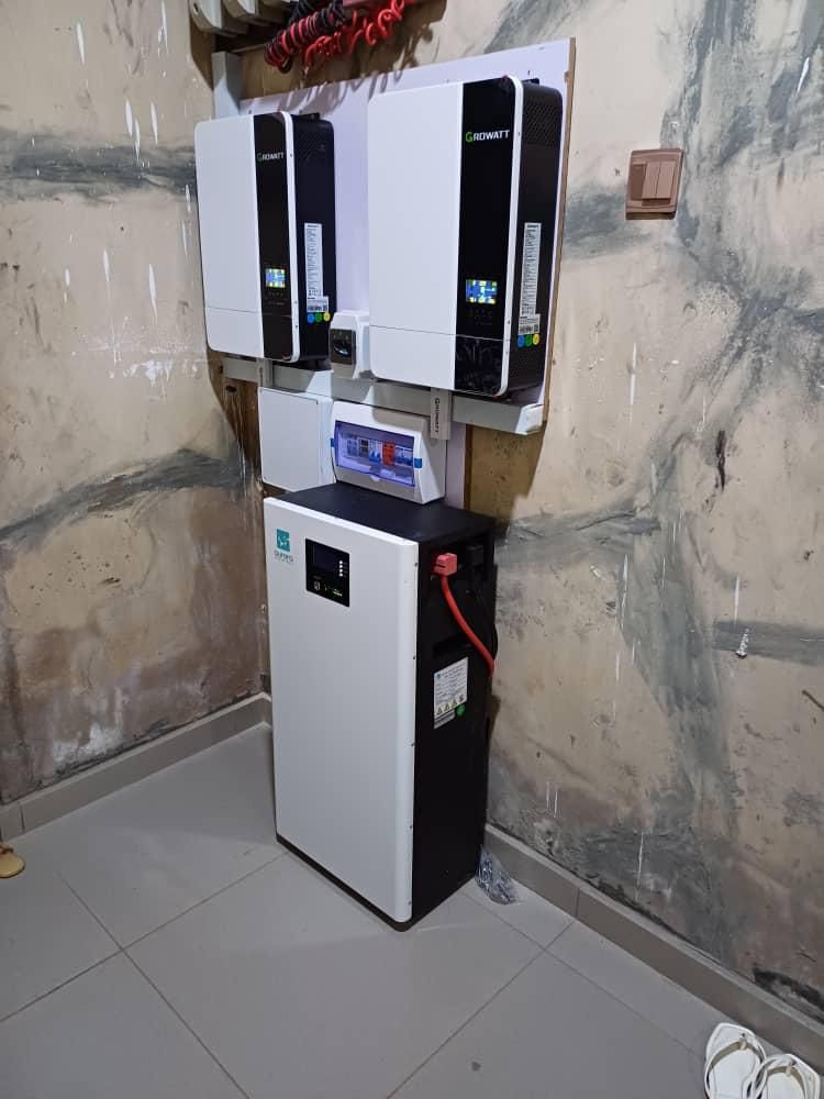 A wall-mounted solar inverter system with connected batteries in a room with tiled flooring and unfinished walls.