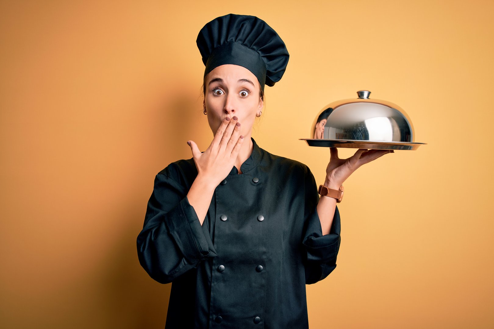 Young beautiful chef woman wearing cooker uniform and hat holding tray with dome cover mouth with hand shocked with shame for mistake, expression of fear, scared in silence, secret concept