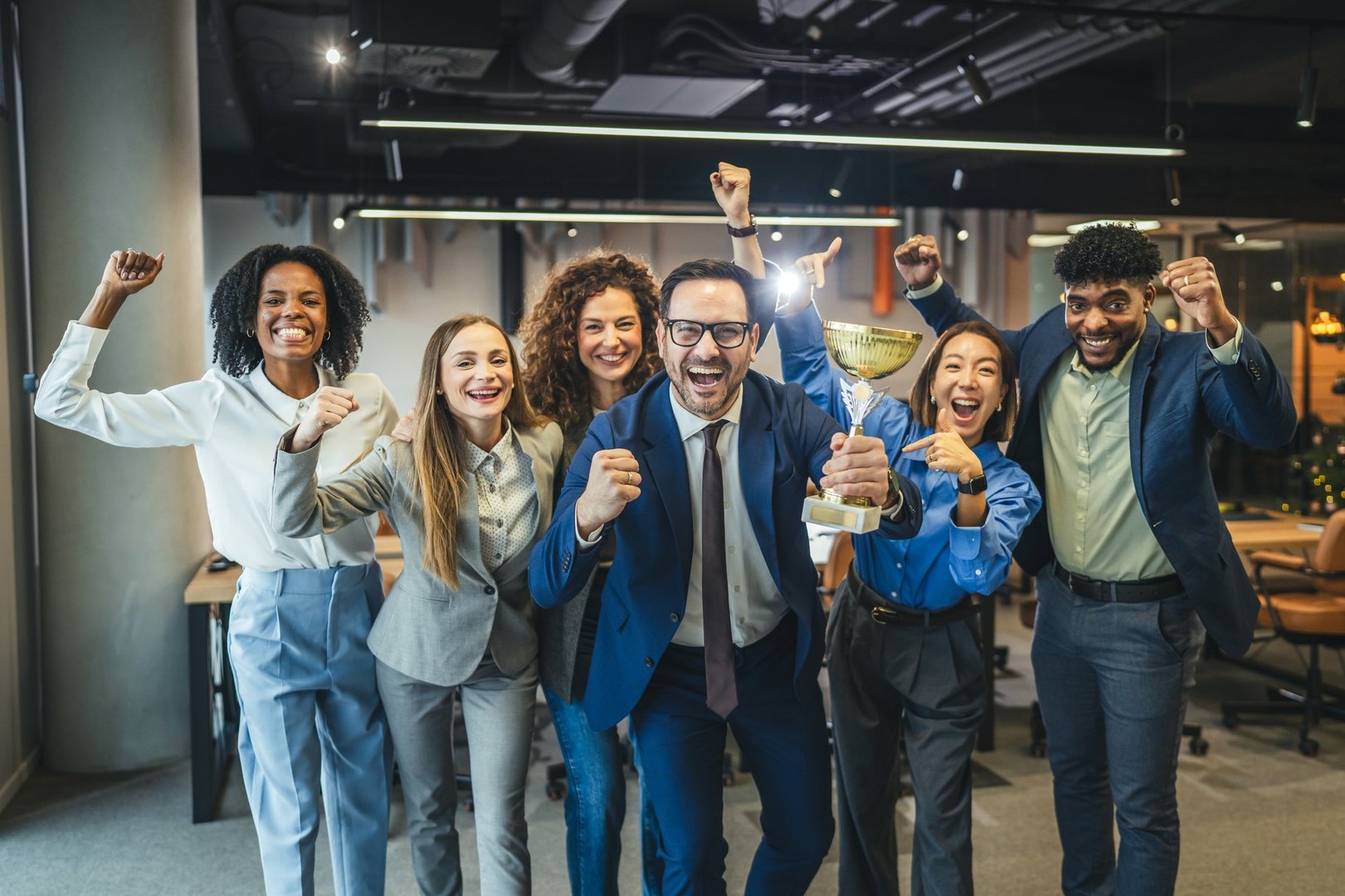 Diverse group of happy business people standing in the office, cheering and celebrating a win, holding a golden trophy, symbolizing teamwork, corporate achievement, and successful collaboration