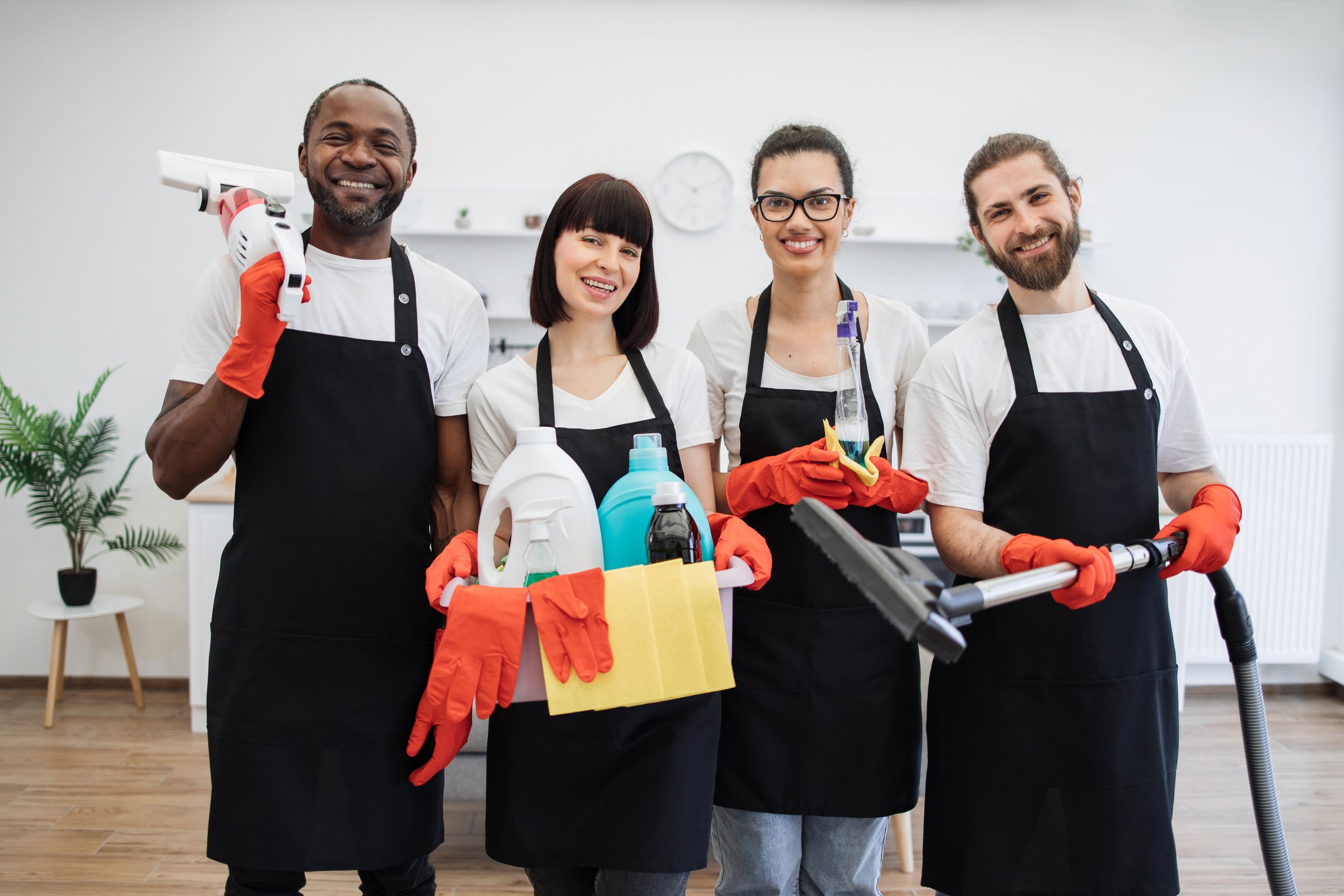 Portrait of happy multiethnic janitors holding cleaning equipment against background of light modern kitchen. Cleaning team, group of four multiracial people in black aprons and red gloves.