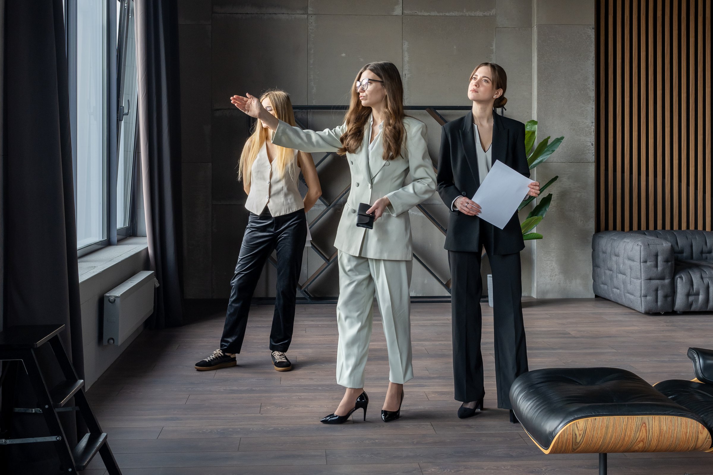 Three business women in office interior, one shows view from window, teamwork during discussion and property presentation.