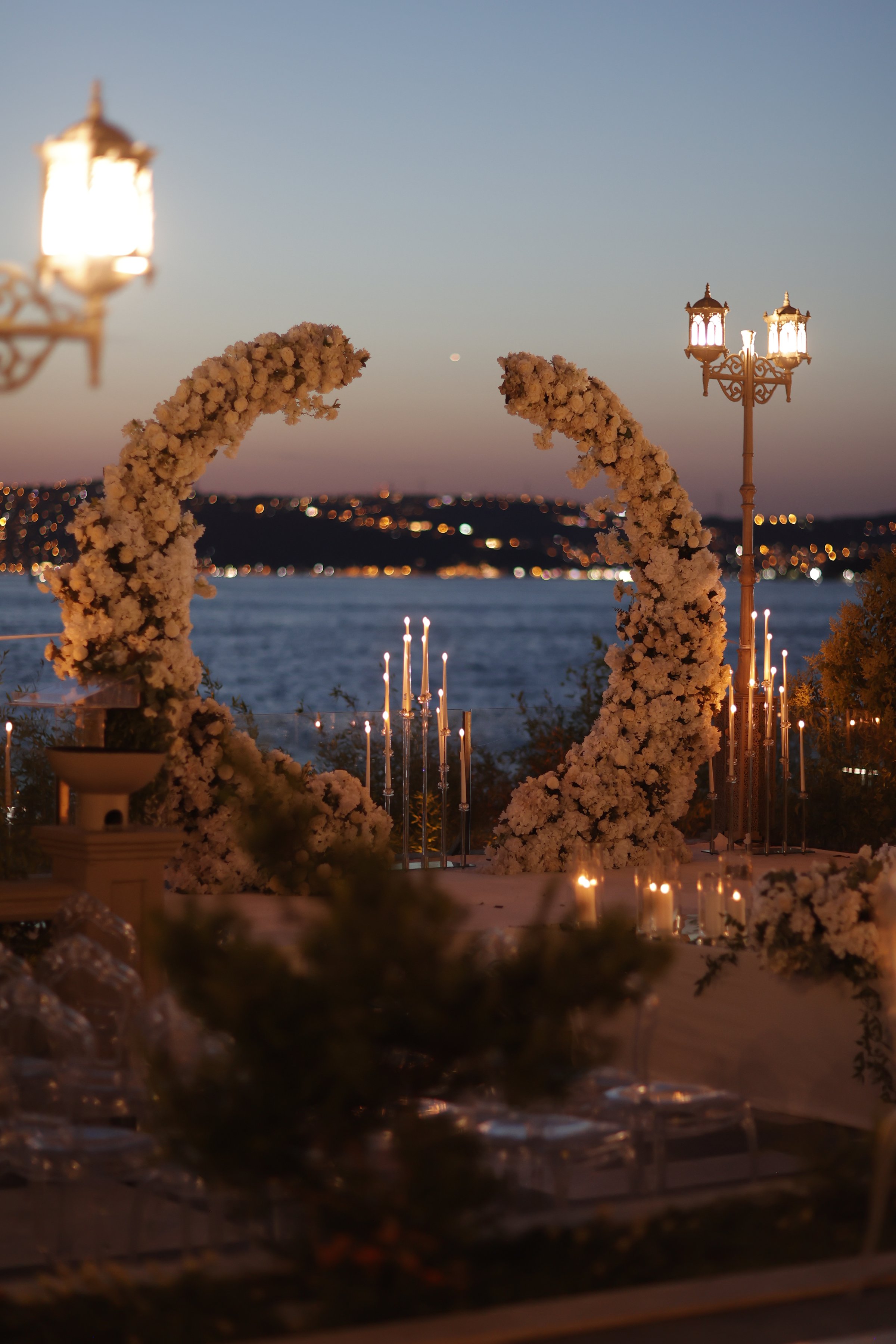 Elegant outdoor wedding ceremony setup at sunset featuring a luxurious crescent-shaped floral arch adorned with white blossoms. Soft candlelight, decorative lanterns, and a stunning Bosphorus sea view create a romantic and sophisticated atmosphere. Perfect for themes of wedding decor, luxury events, romance, celebration, and destination weddings.