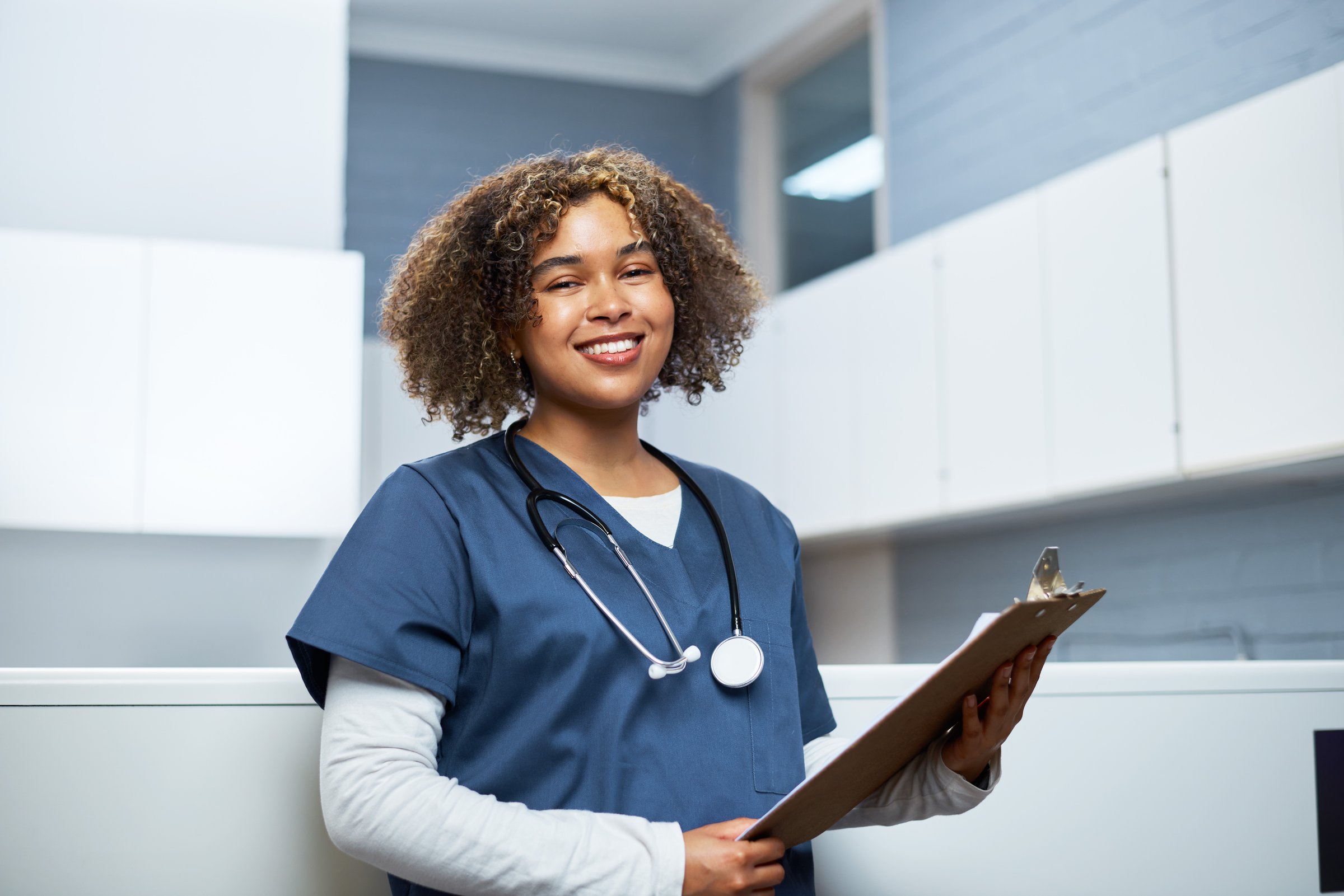 Young healthcare provider in scrubs standing confidently in a well-lit medical facility, holding a clipboard and wearing a stethoscope, symbolizing care, professionalism, and a welcoming atmosphere in medical environments.