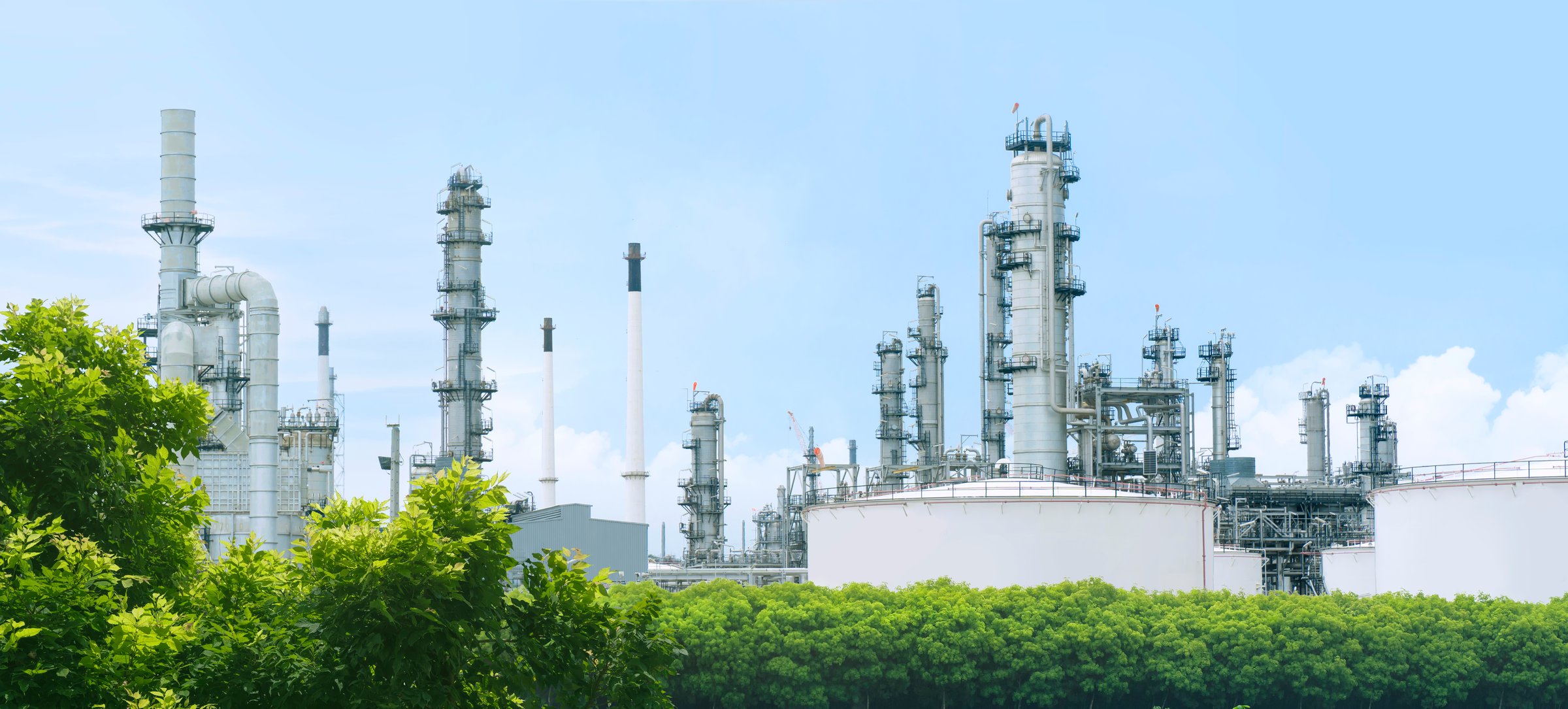 Oil refinery plant from industry zone with lush green trees and blue sky, large Oil and gas petrochemical industrial, Refinery factory oil storage tank and pipeline, Ecosystem and good environment.