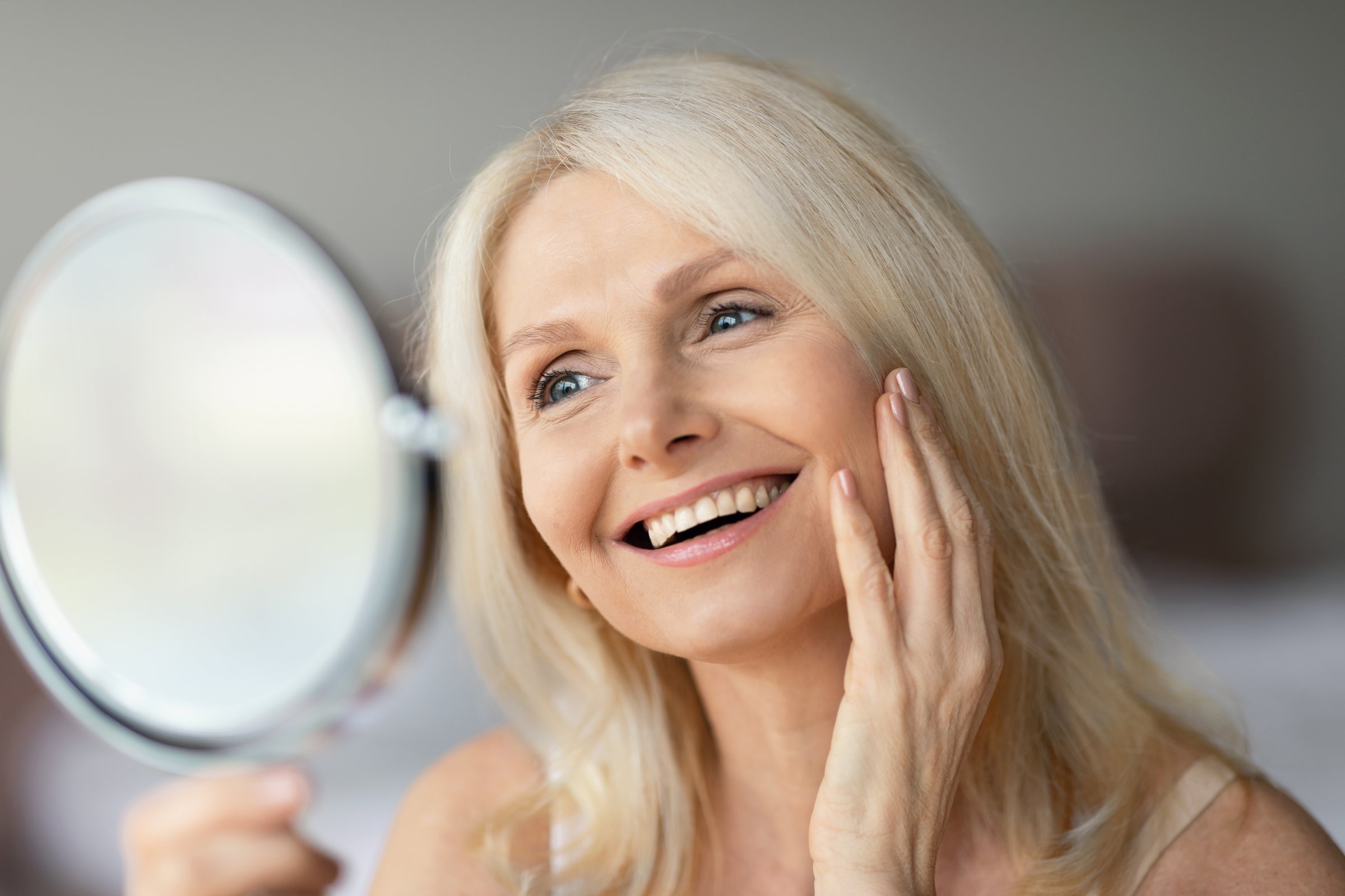 Portrait of beautiful senior woman looking in miror and touching face, checking her wrinkles and smiling, admiring her beauty. Mature lady taking care of her skin, doing anti-aging procedures