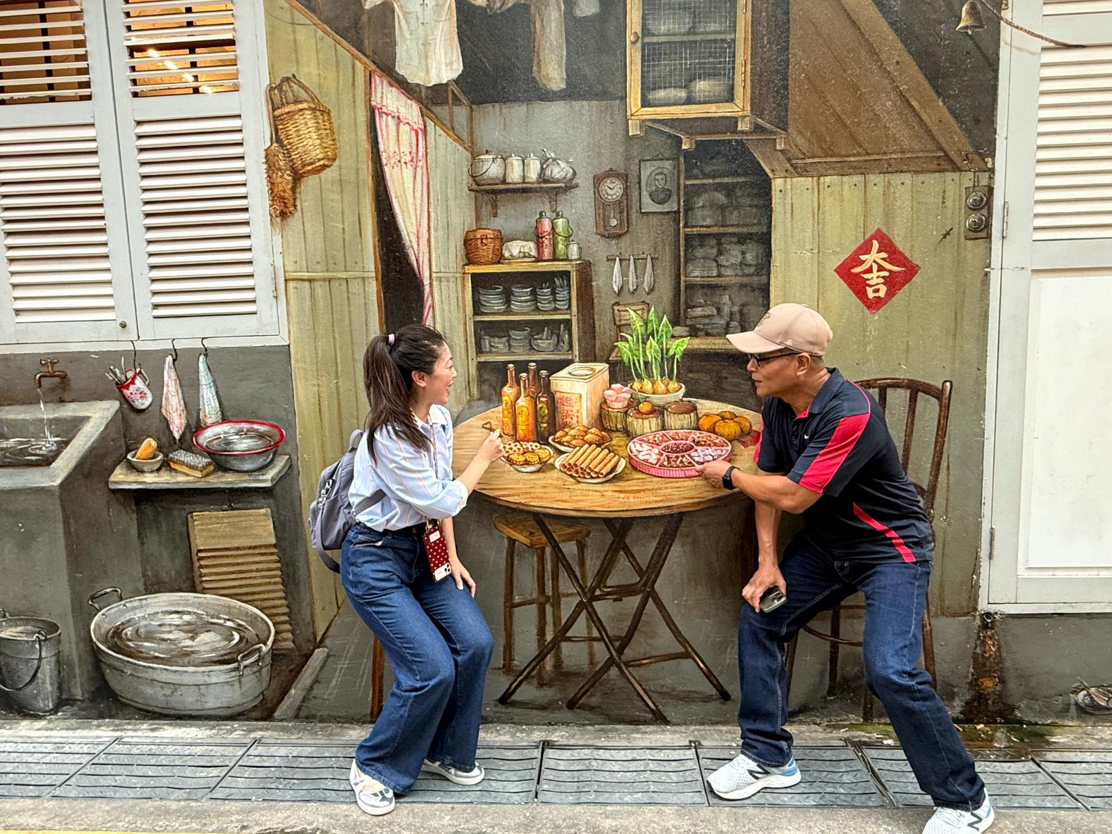 Two people pose playfully in front of a realistic wall mural depicting a vintage kitchen scene with various food items.