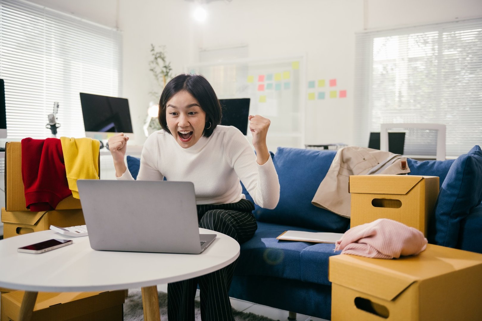 Young asian businesswoman is sitting on a comfortable blue sofa in her new office. Surrounded by cardboard boxes. Raising her arms in excitement as she achieves success while working on her laptop