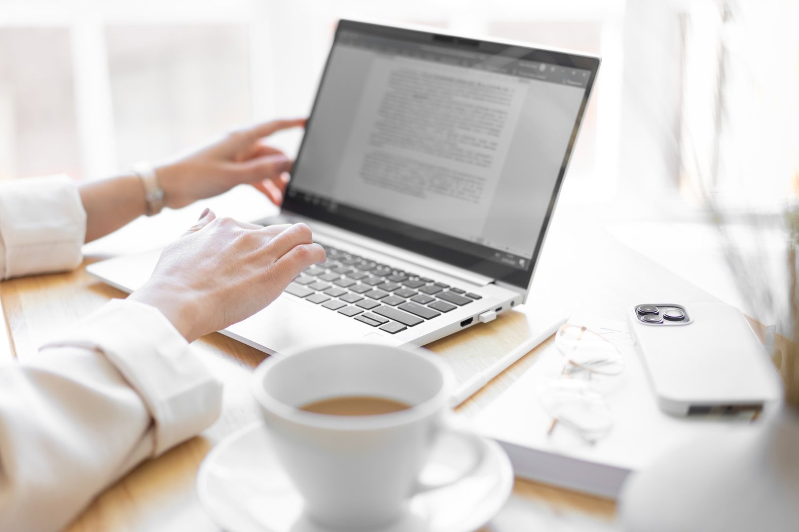 Close-up of the right female hand. Unrecognizable woman is typing text on a laptop. Cup of coffee, smartphone, glasses on the desktop. Blurred foreground.