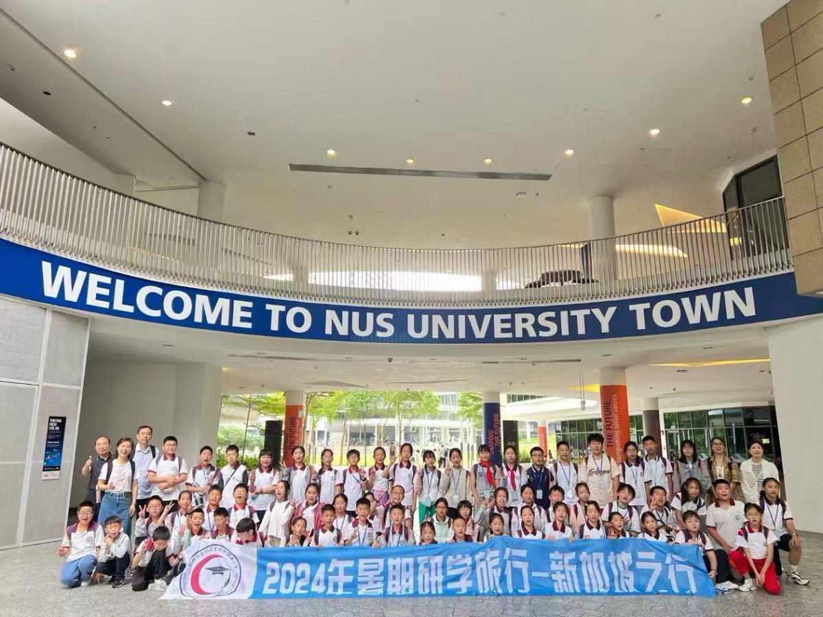 A group of children and adults pose with a banner under a Welcome to NUS University Town sign.
