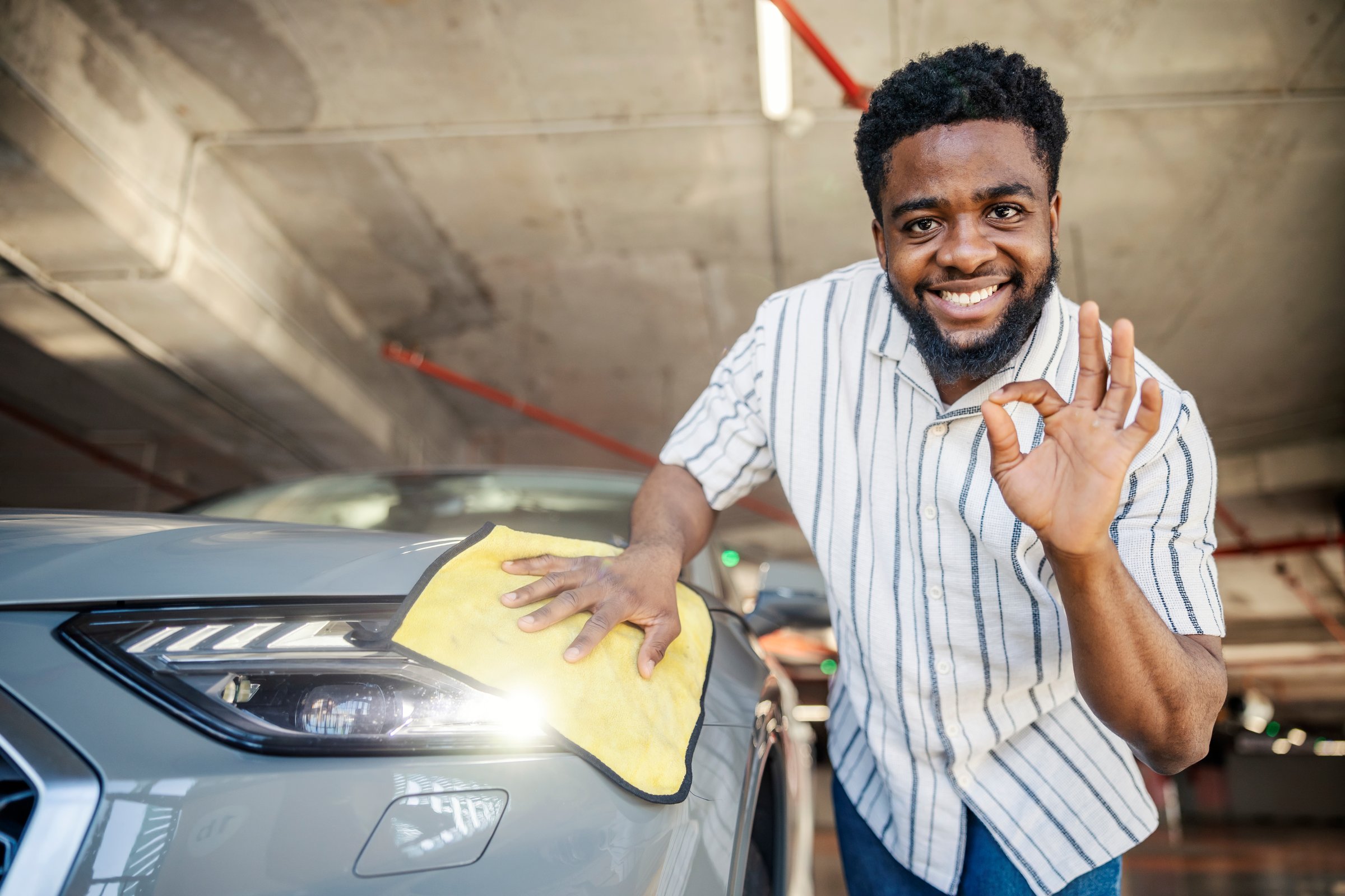 Smiling young black man is gesturing okay while cleaning headlights on his car at public garage.