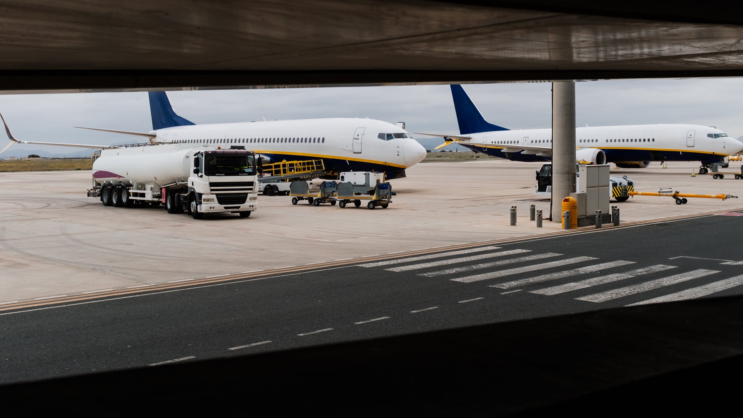 Tanker truck refueling airplane on airport tarmac with luggage carts waiting nearby