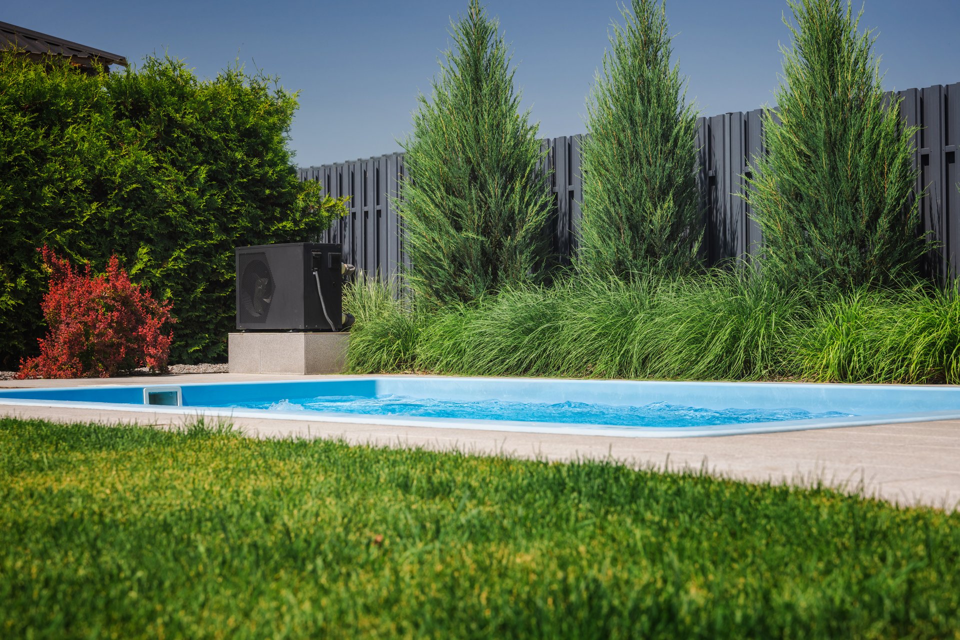 Modern heat pump installed next to a backyard pool, used for heating water, surrounded by green lawn, conifers, and privacy fence in a landscaped garden.