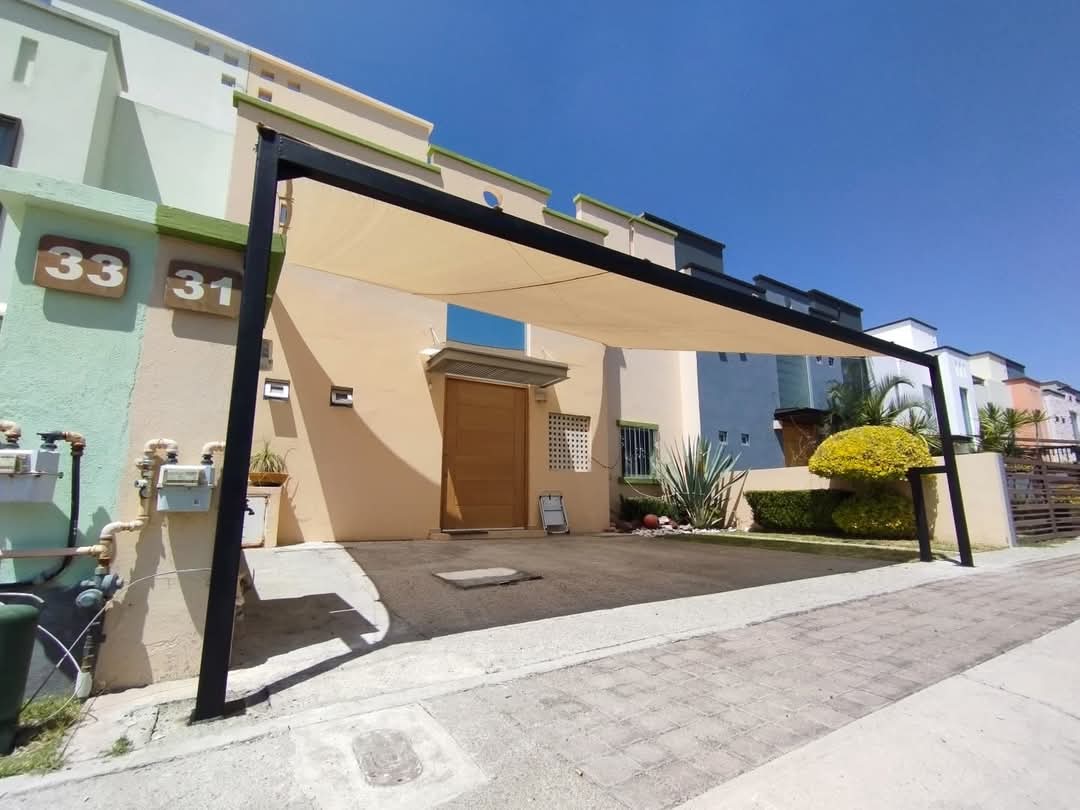 Modern house exterior with a triangular shade canopy over a parking space, surrounded by plants and a clear blue sky.