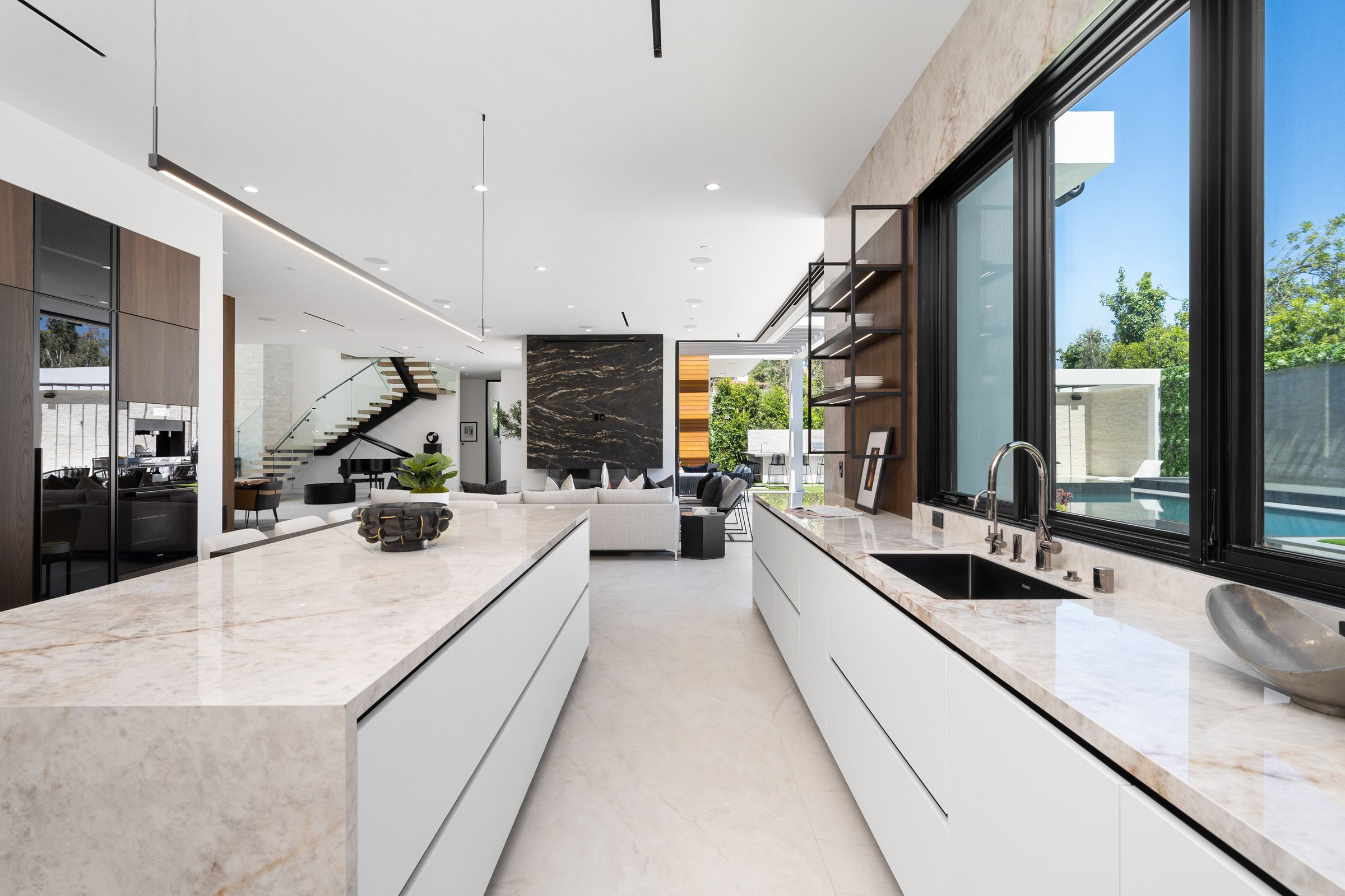 Modern kitchen interior with marble countertops, large windows, and a view of a living area and staircase.