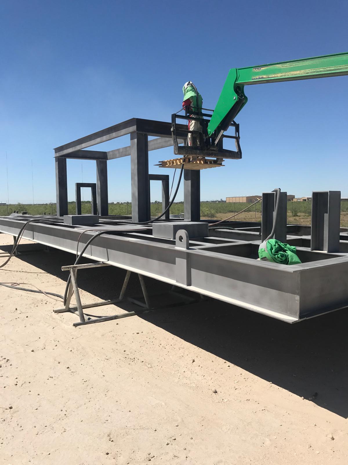 A worker on a green lift machine assembling a large metal structure outdoors against a clear blue sky.