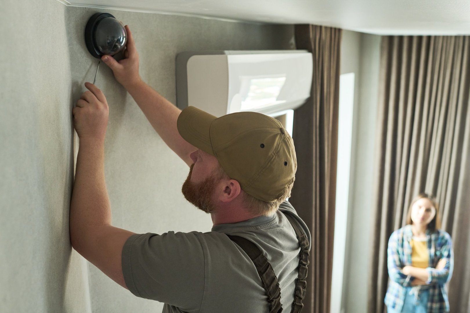 Caucasian middle aged man installing security camera on wall while young Caucasian woman standing in background observing process in modern residential interior