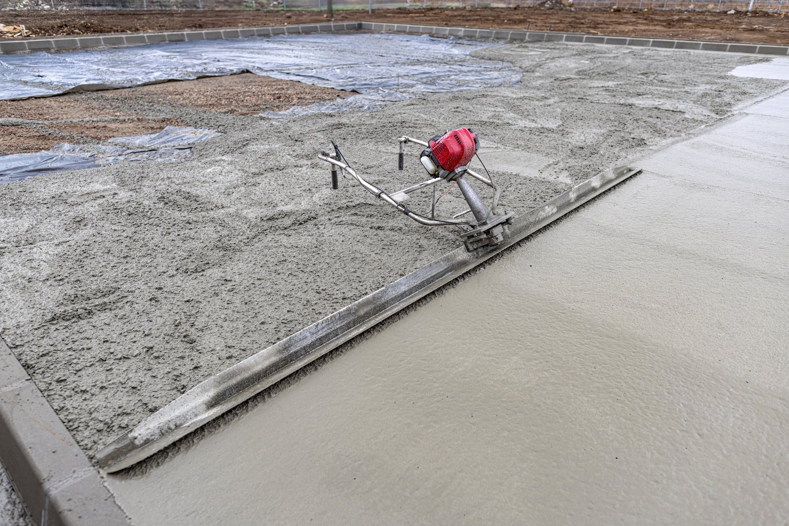 Power screed smoothing fresh concrete surface at construction site with grader in background