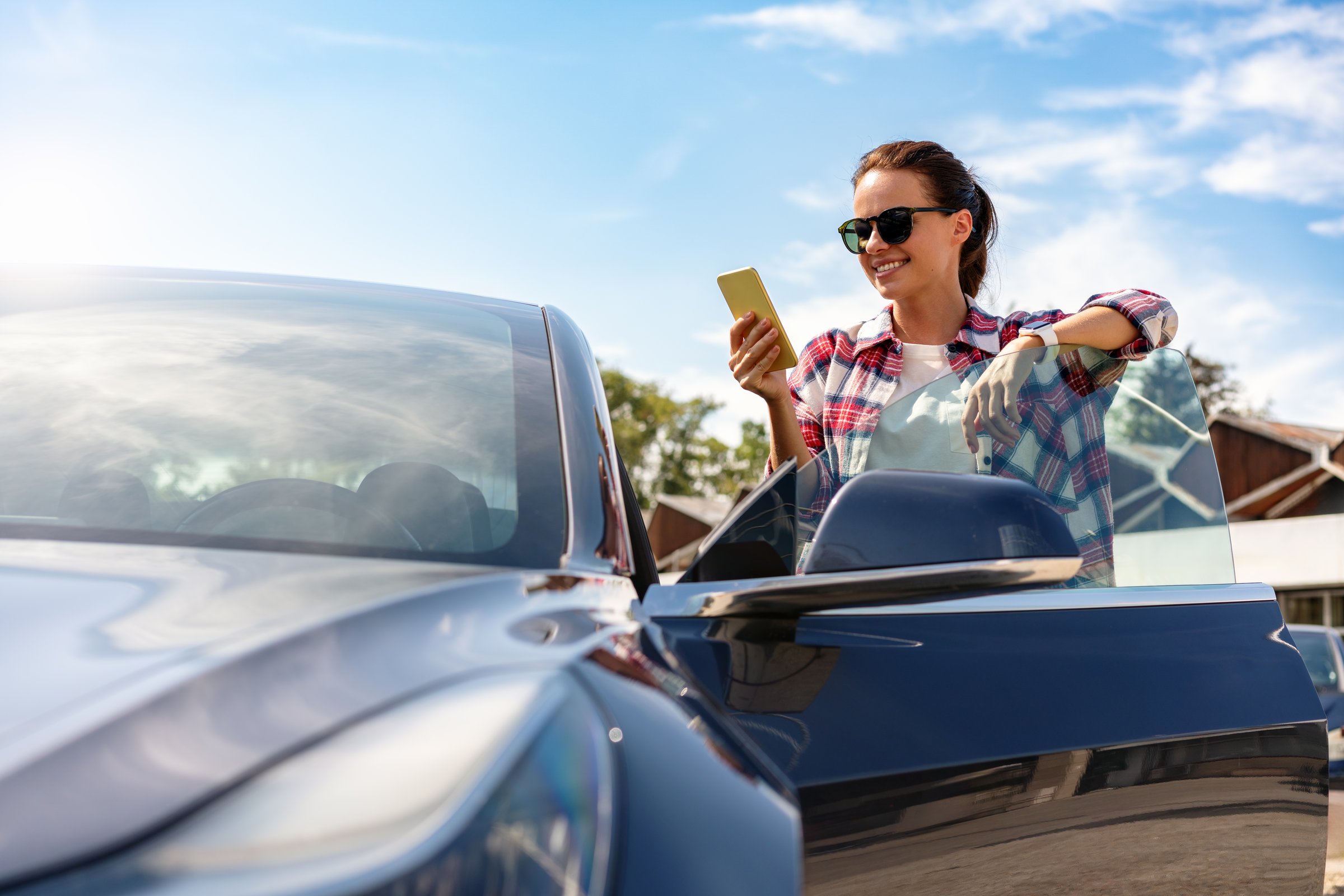 Woman looking at screen of mobile phone while standing next to her car outdoors