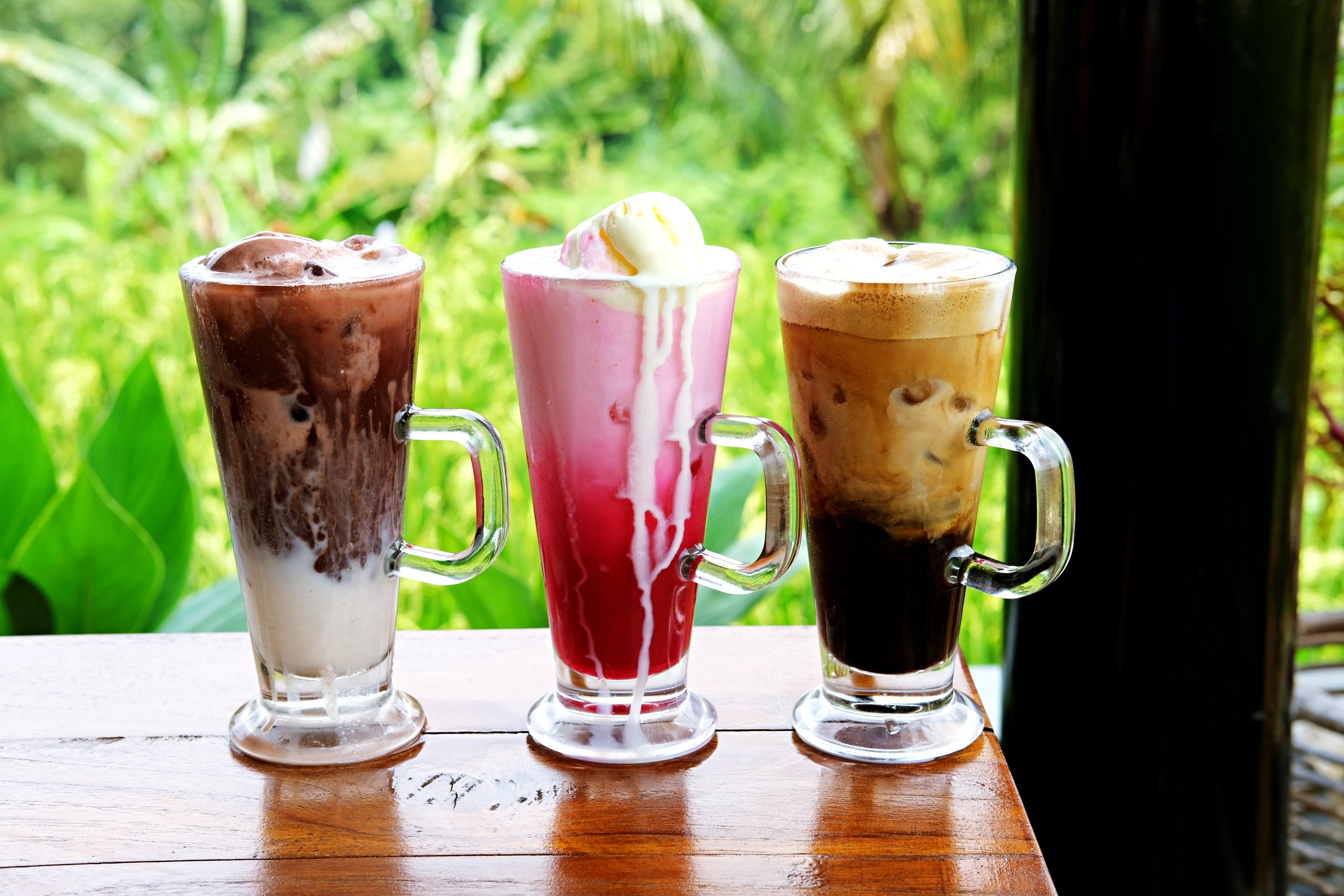 Three colorful iced drinks served in tall glasses on a wooden table, with a fresh green garden background. Perfect for beverage, summer refreshment, cafe menu, and lifestyle concepts.