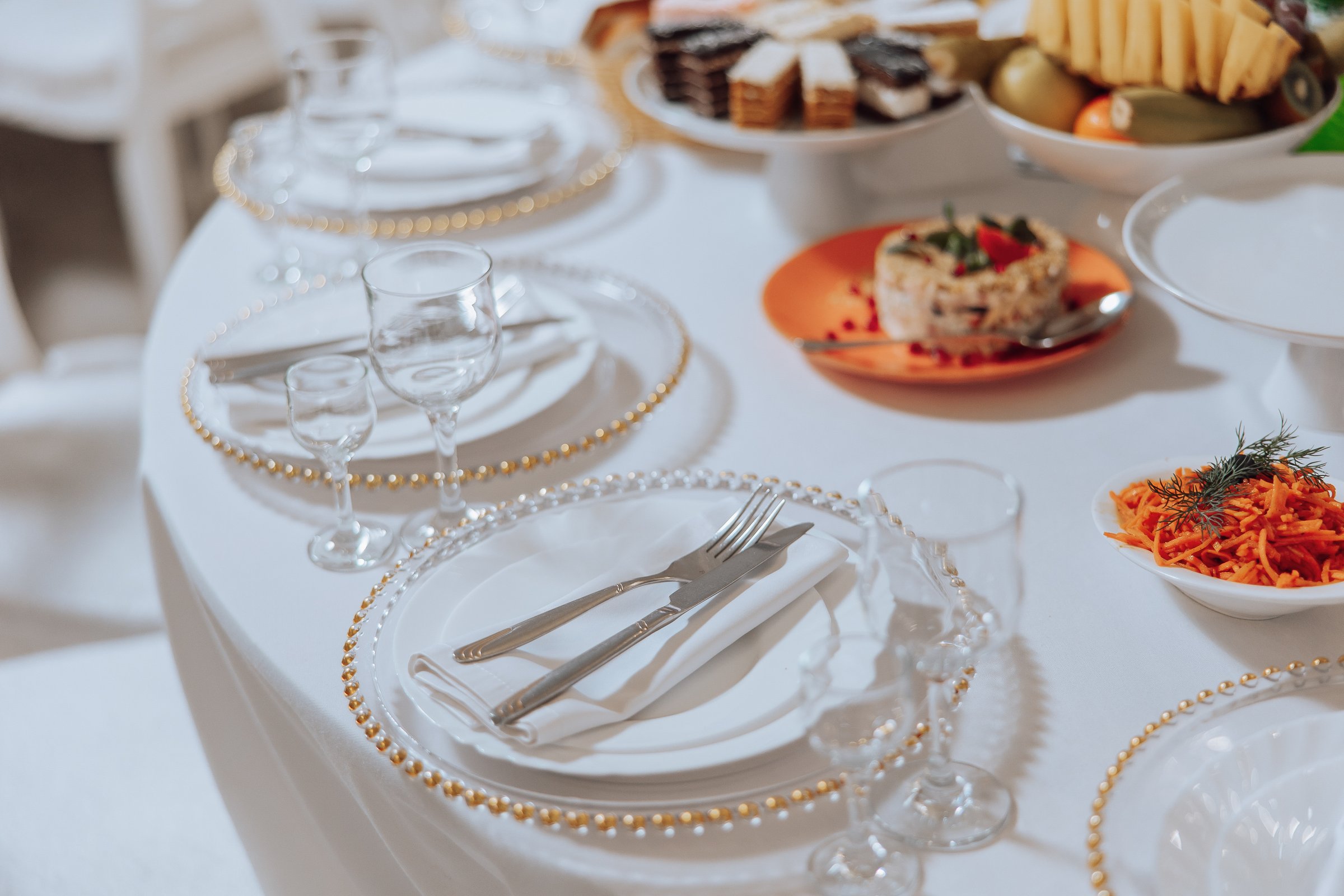 A table with a white tablecloth and a variety of food and drinks. The table is set with silverware and glasses, and there are several plates of food on the table. Scene is elegant and inviting