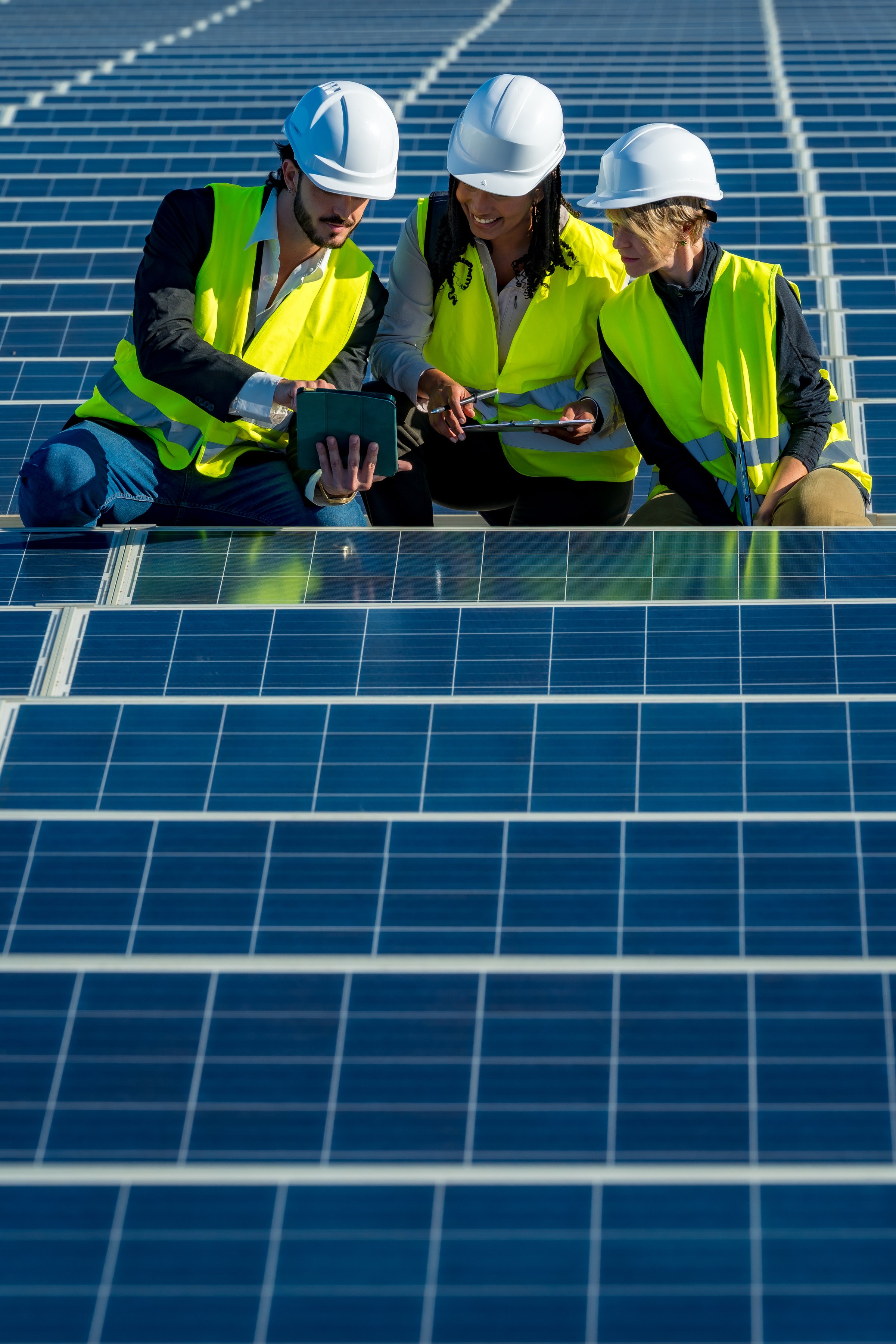 Group of engineers wearing hard hats and safety vests discussing data on a digital tablet while performing an inspection of solar panels at a renewable energy farm