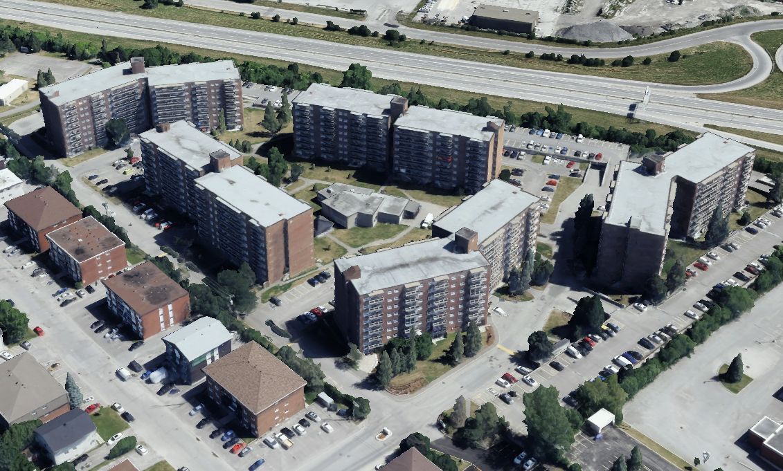 Aerial view of several large apartment buildings with parking spaces, surrounded by roads and greenery.