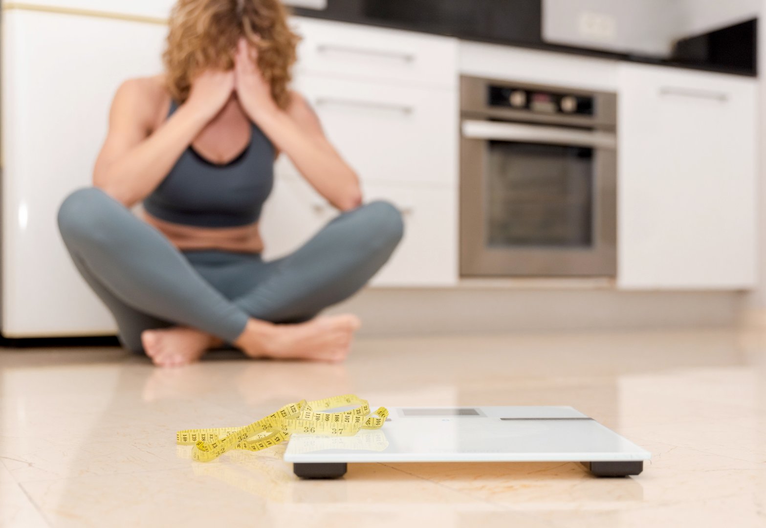 Upset woman in sportswear sitting on kitchen floor with scale and measuring tape, weight loss struggle